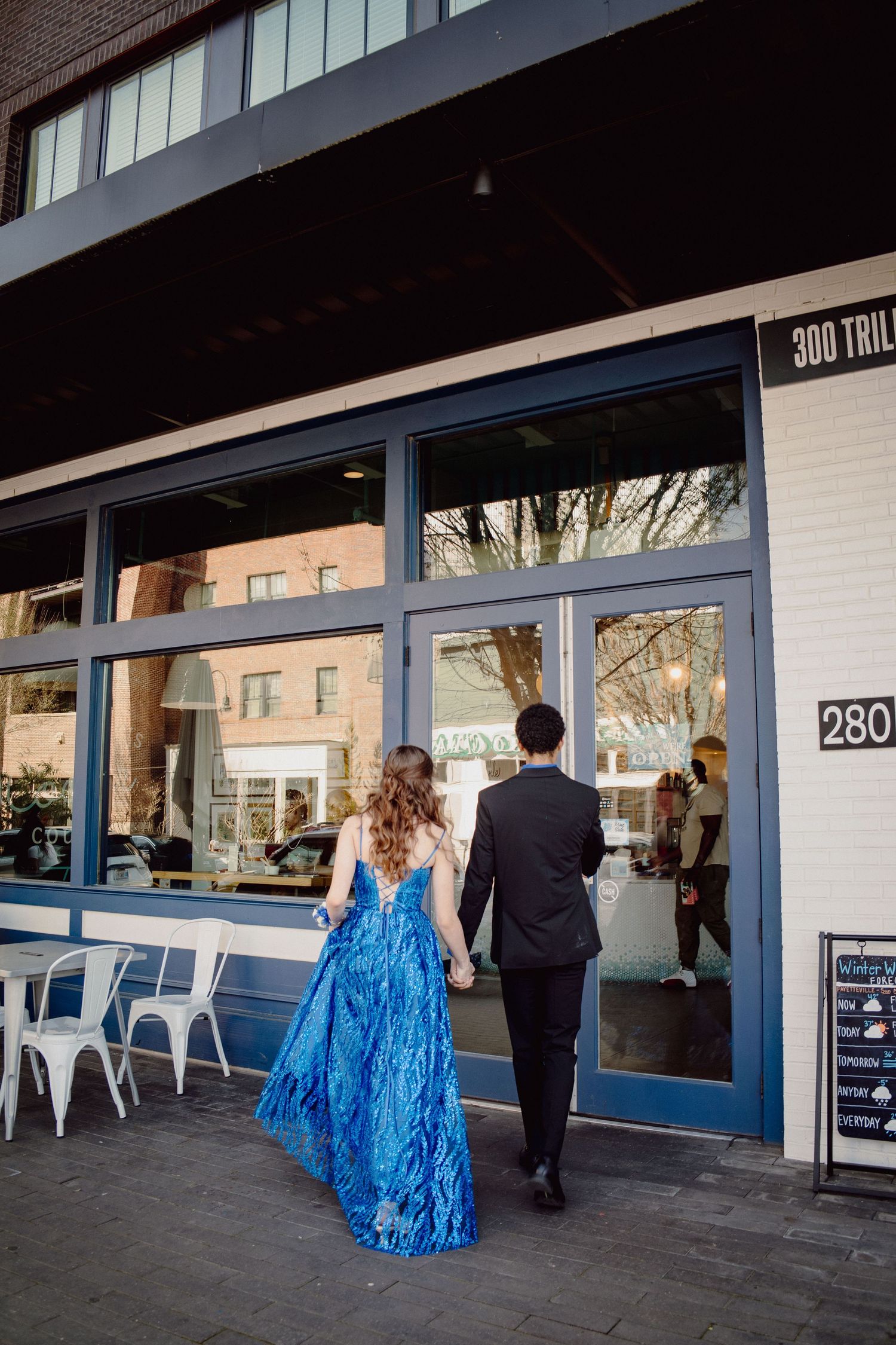 A prom couple holds hands walking past a modern storefront, the woman in a flowing blue sequined gown.