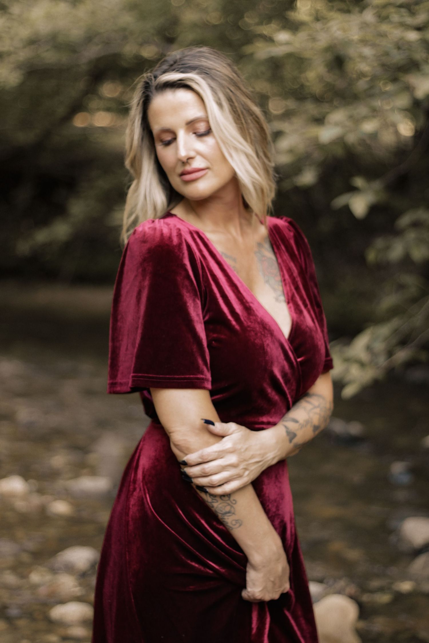 Woman wearing red velvet wrap dress poses outdoors against blurred forest backdrop.