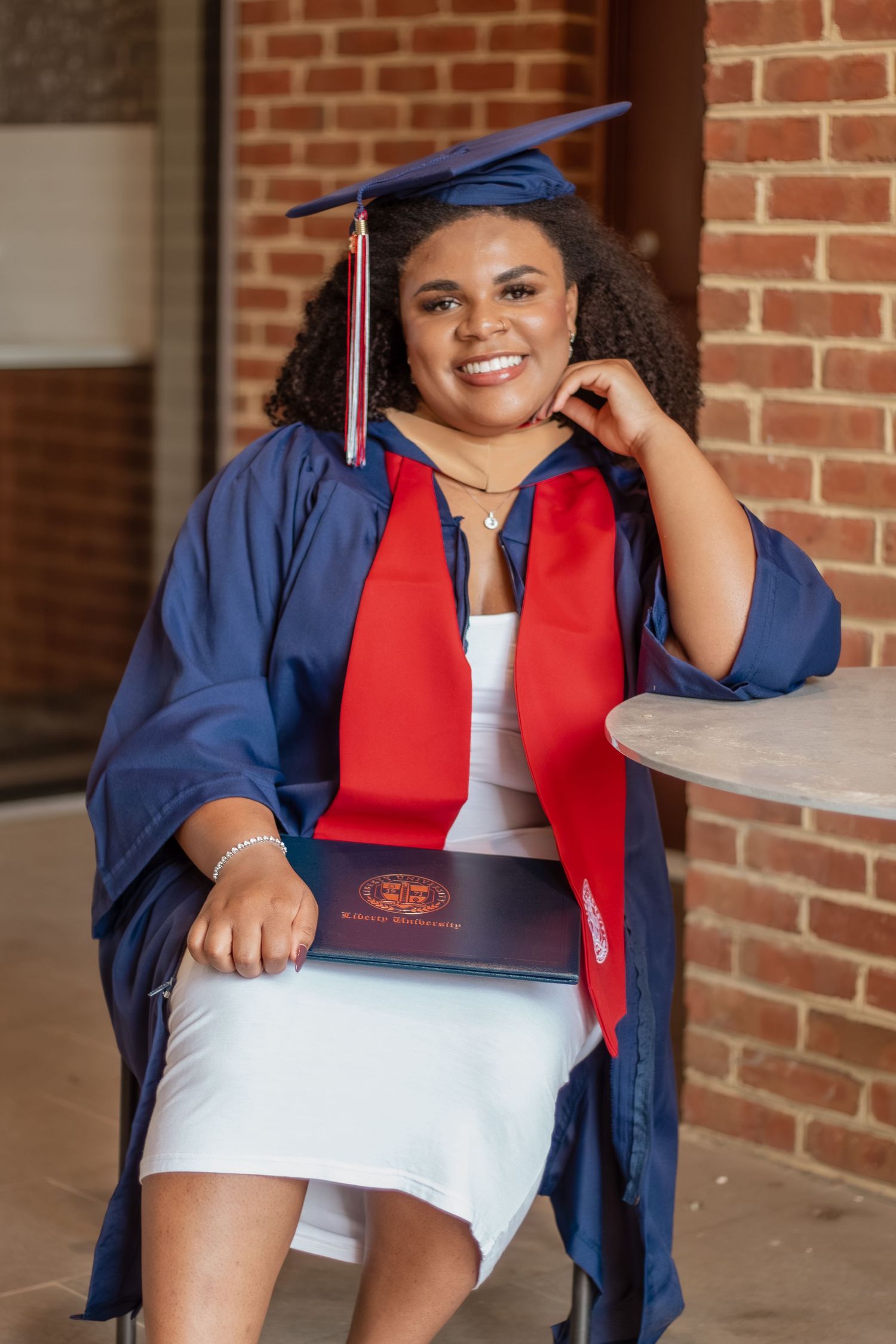 Graduate posing with diploma in blue cap and gown against brick background.