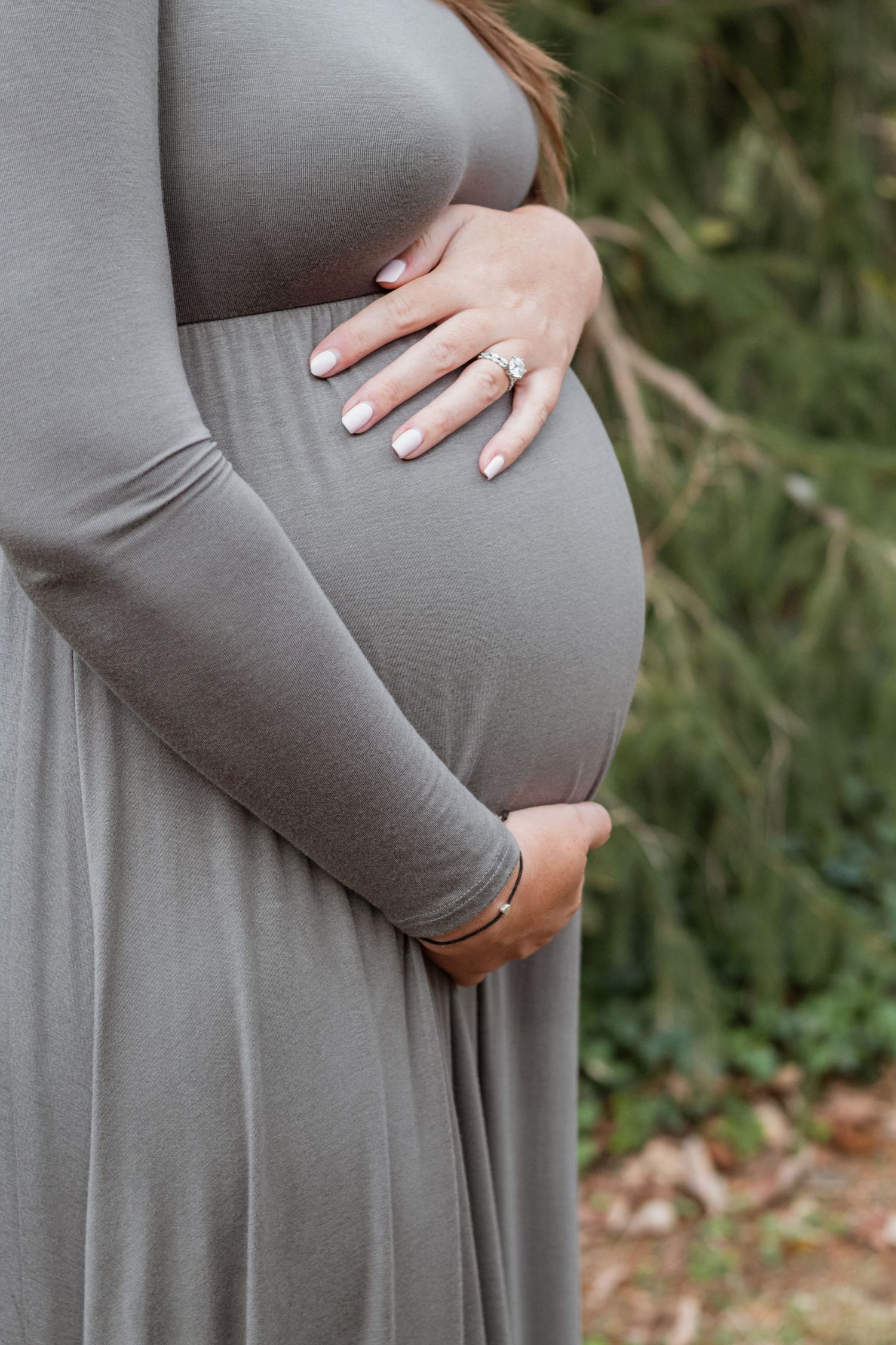Close-up of pregnant belly with hands showing engagement ring in a grey maternity dress.