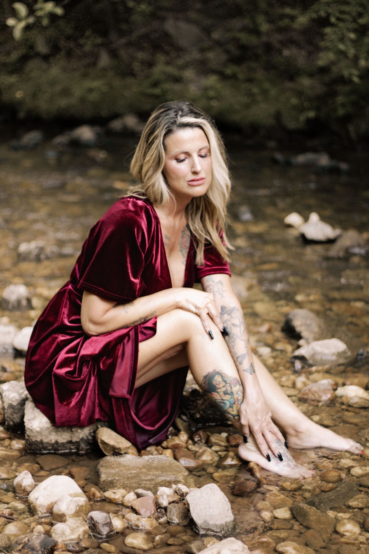A person in a deep red velvet dress sits on rocks by a stream.