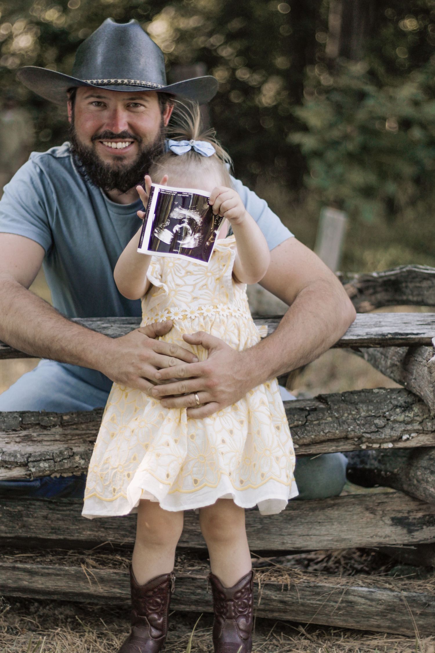 Person in cowboy hat holding small child displaying ultrasound photo on rustic fence.
