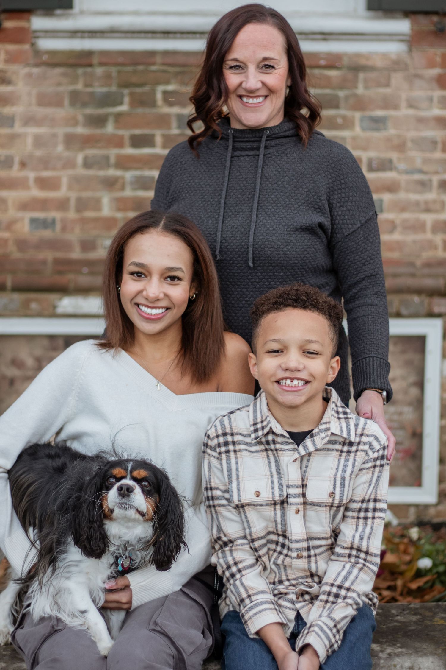 Family portrait session featuring a small dog, plaid shirt, and brick wall backdrop.