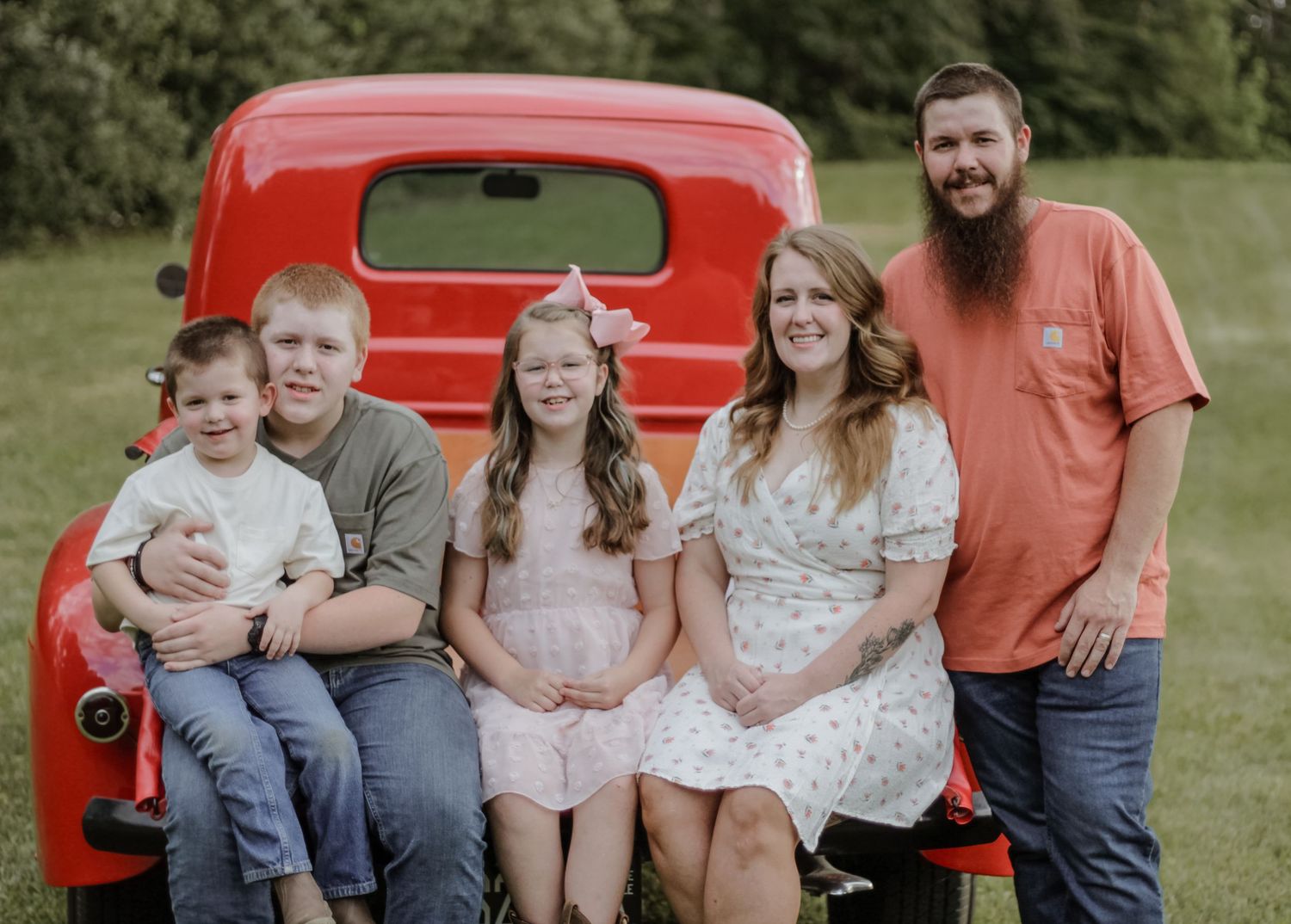 A family sits together on the back of a vintage red pickup truck in an outdoor setting.