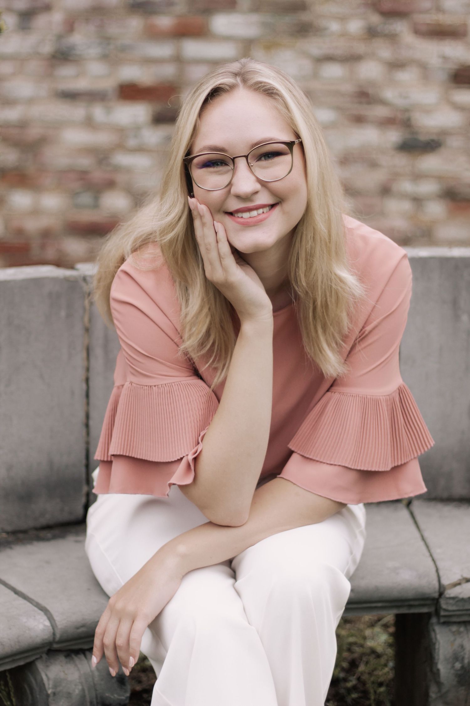 Person in pink ruffled top and white pants poses against brick wall.