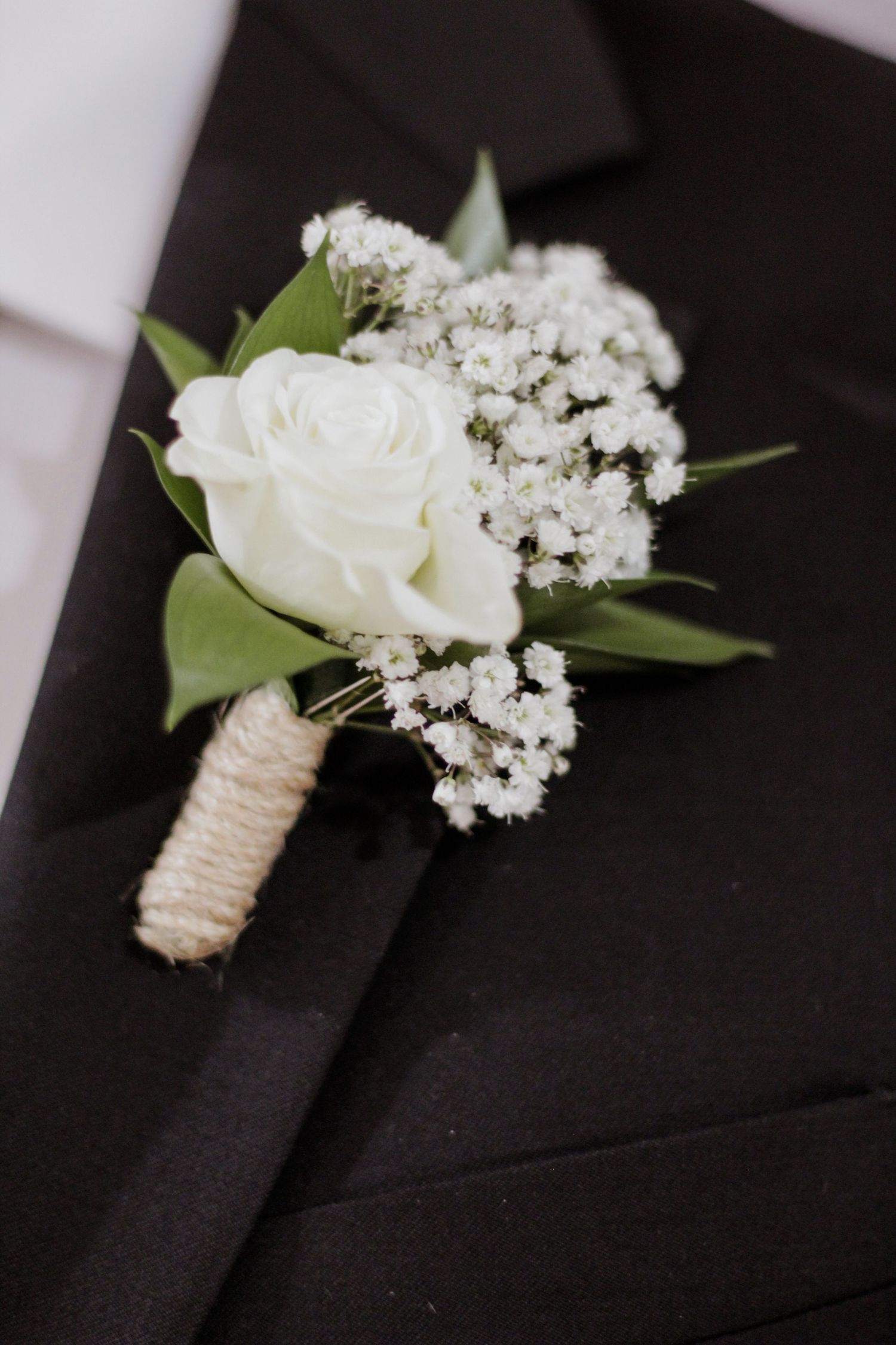 White rose and baby's breath boutonniere with twine wrap on black suit lapel.