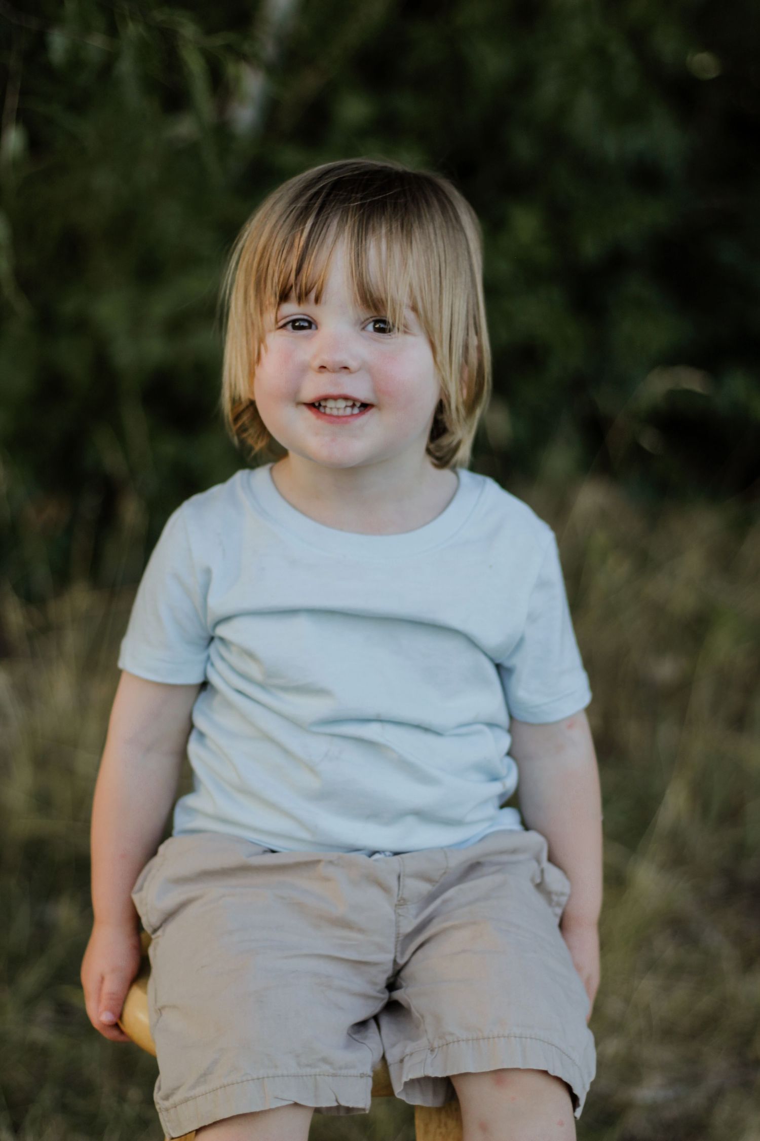 Child wearing light colored t-shirt and shorts smiles while sitting outdoors.