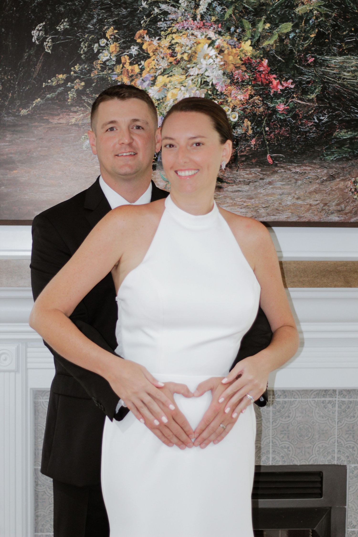 Wedding couple embraces in front of floral backdrop wearing classic formal attire.