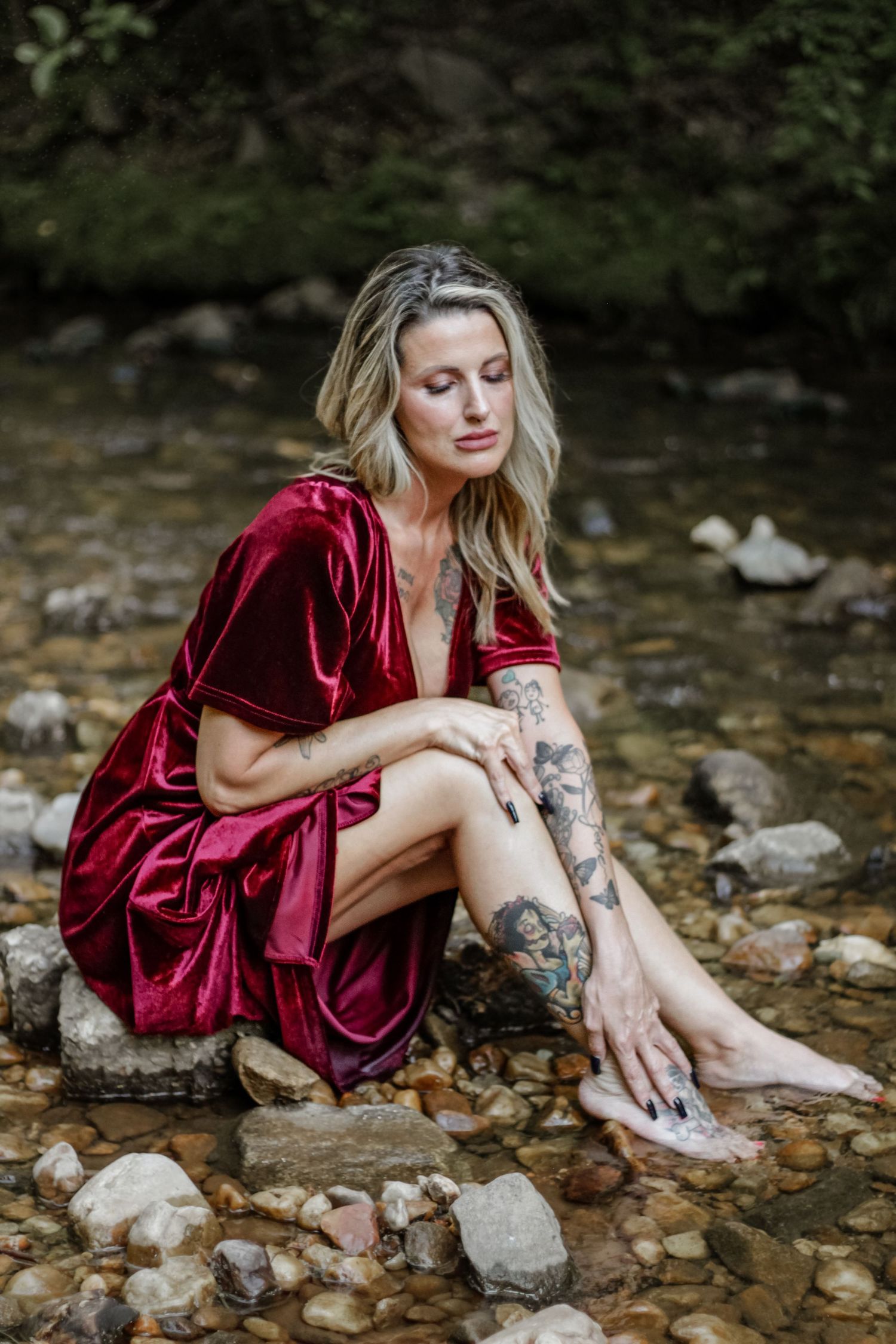 Artistic portraits by stream showing burgundy velvet dress and natural setting.