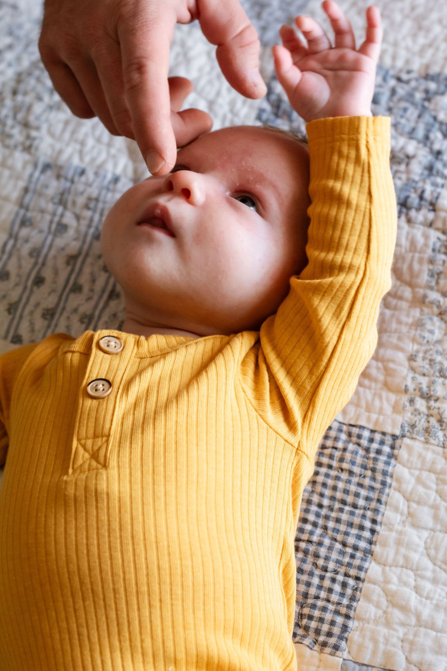 Baby wearing yellow ribbed onesie stretches and yawns while lying on a patterned quilt.