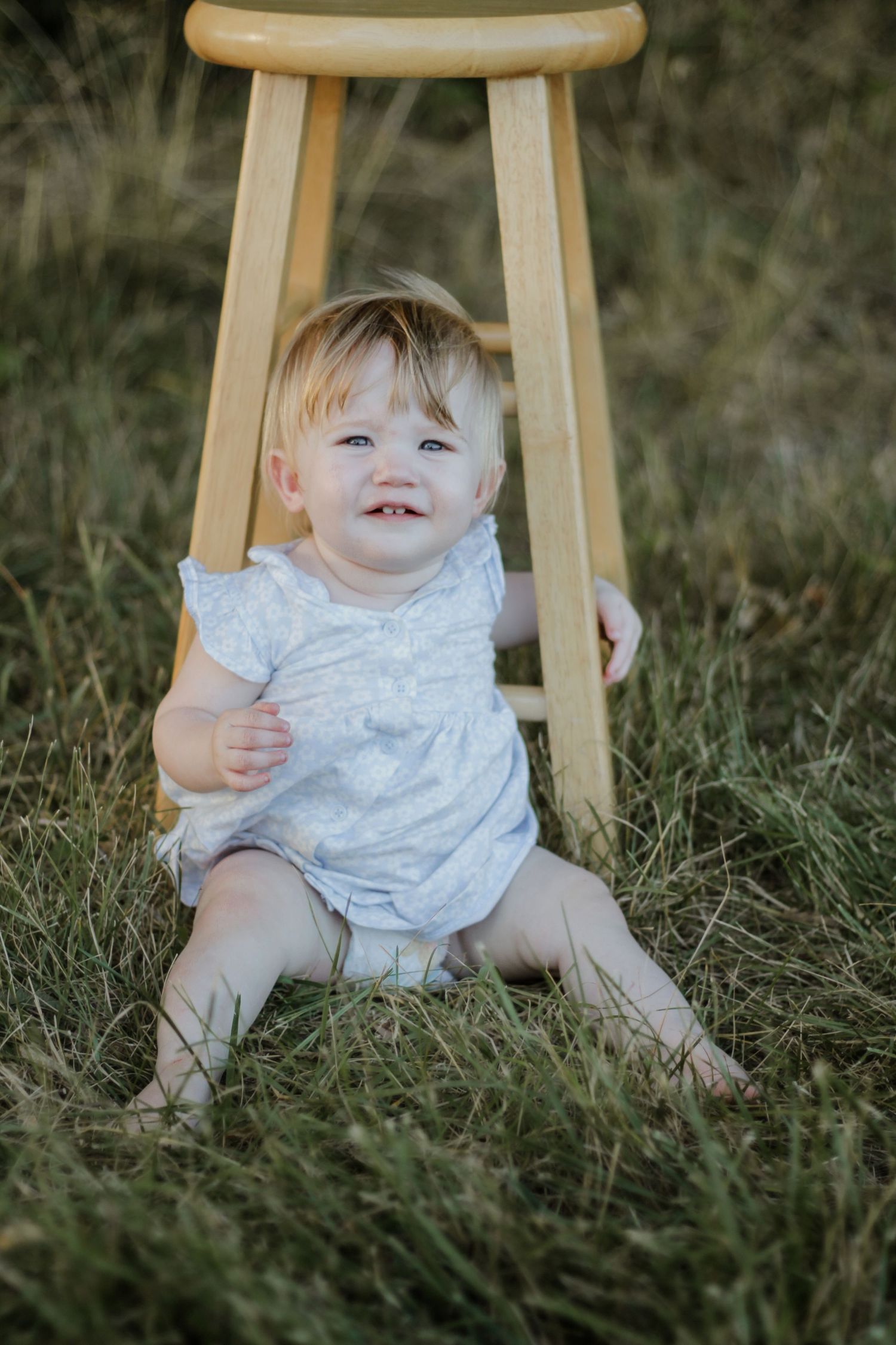 Small child sits near wooden stool in grassy field during outdoor photoshoot.