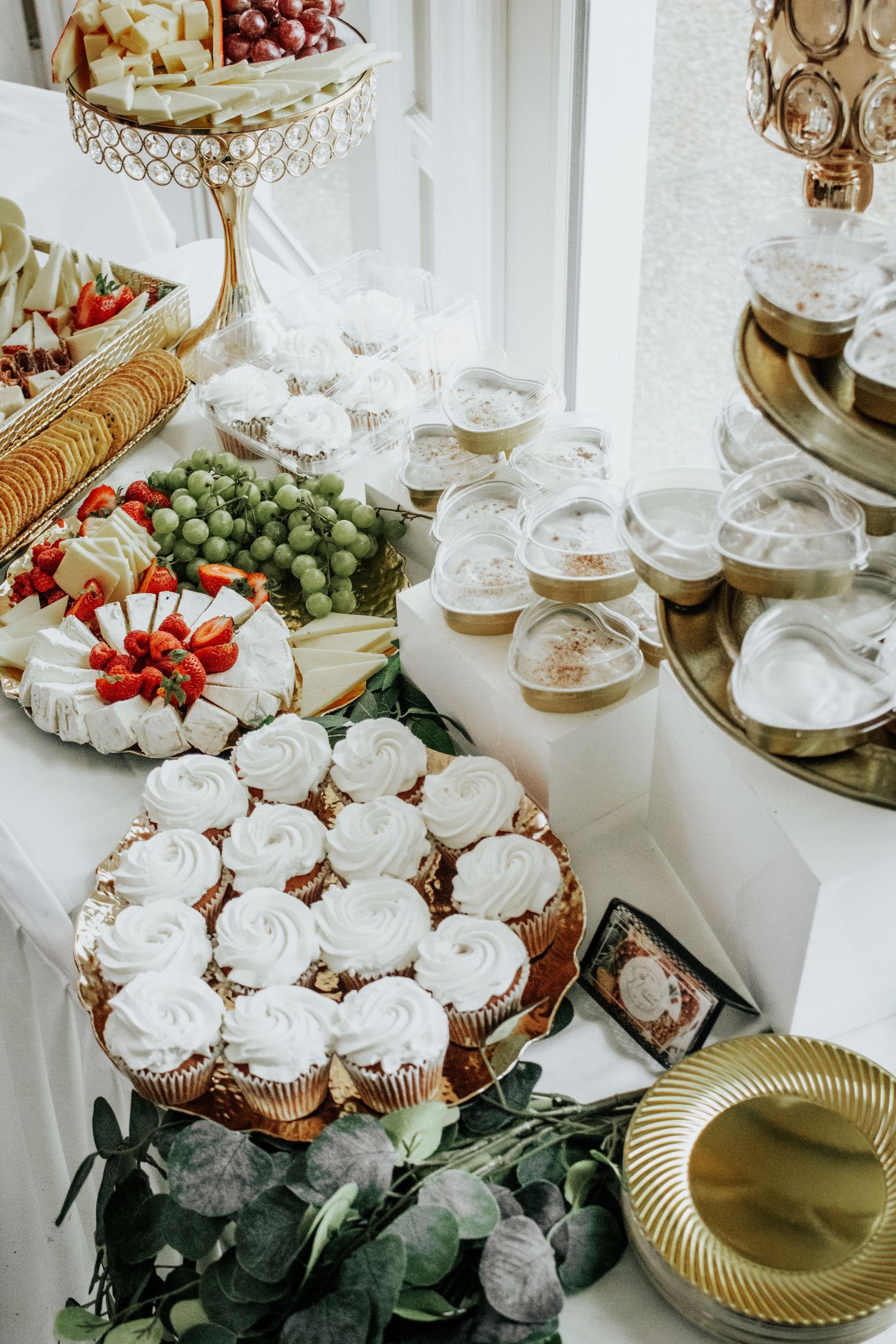 Elegant dessert and appetizer display featuring cupcakes, cheese boards and fresh fruit on tiered stands.