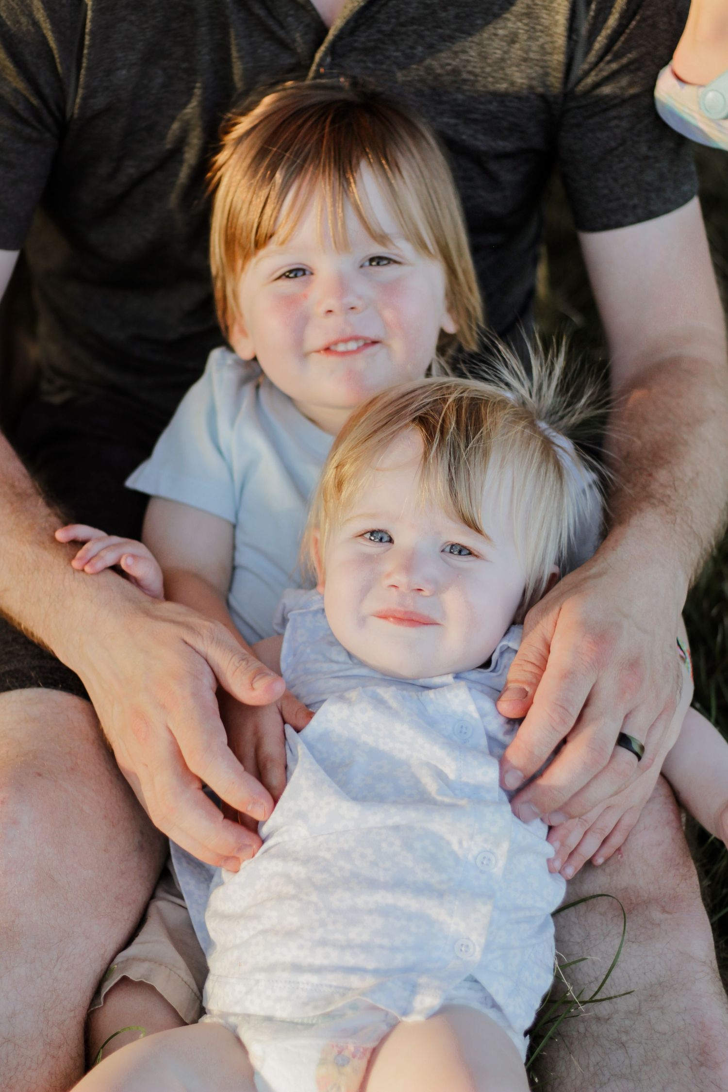 Two young children sitting together on an adult's lap outdoors during a family photoshoot.
