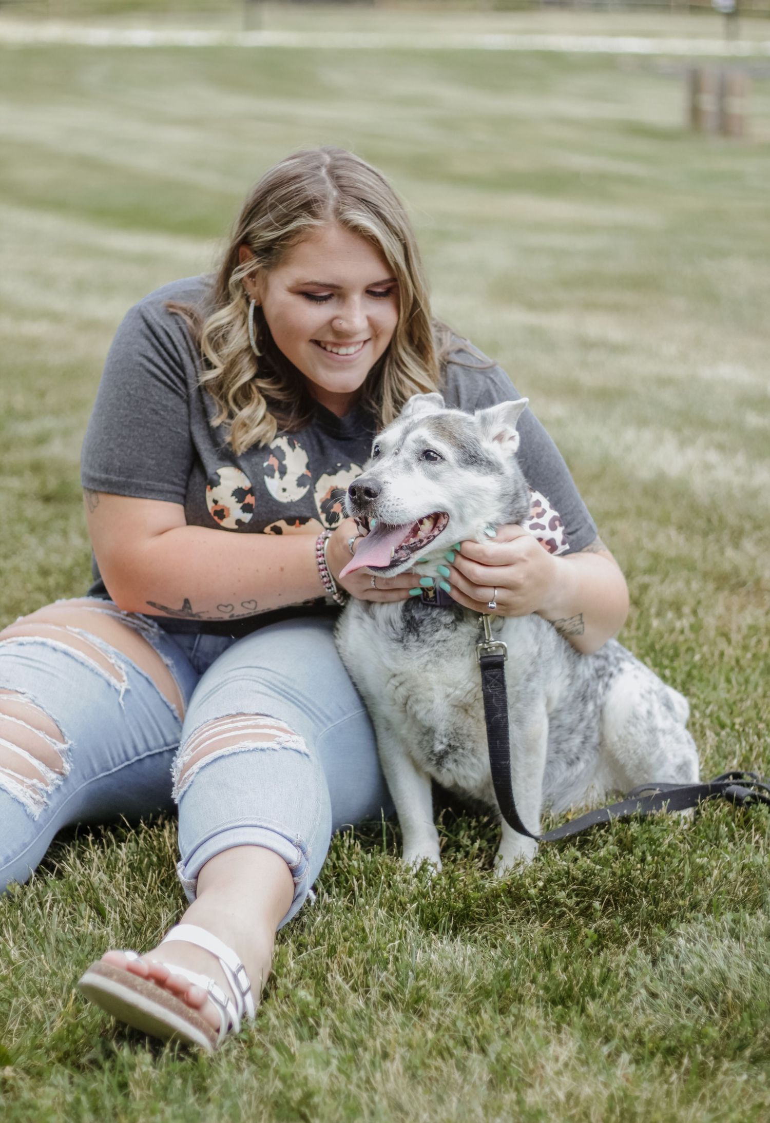 A woman in ripped jeans sits on grass with her happy white dogs.
