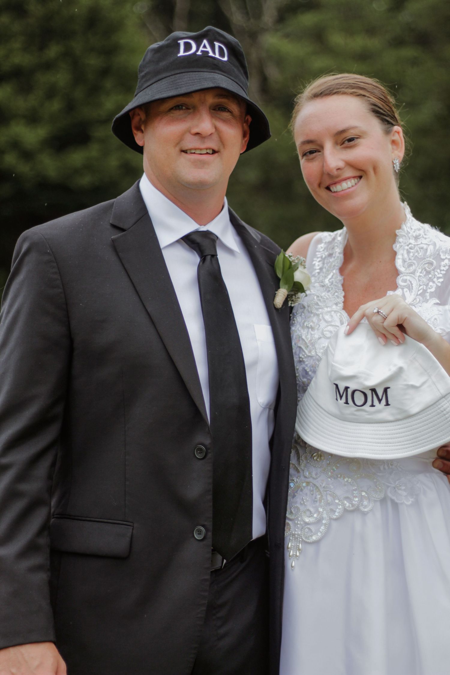 Wedding couple wearing hats labeled 'Mom' and 'Dad' posing together outdoors.
