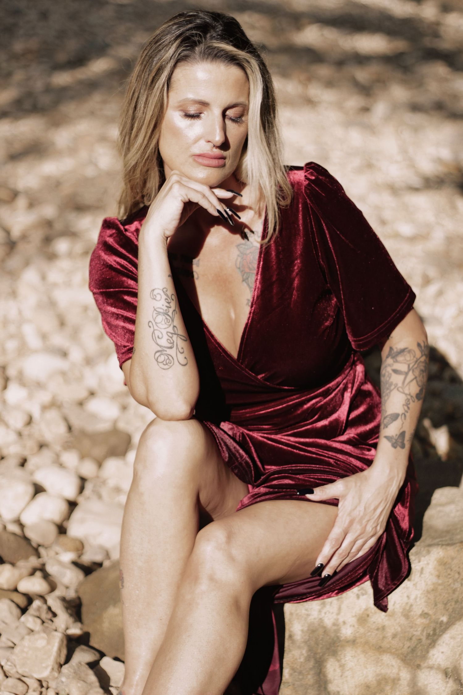 A person in a burgundy velvet dress poses thoughtfully on rocky terrain.