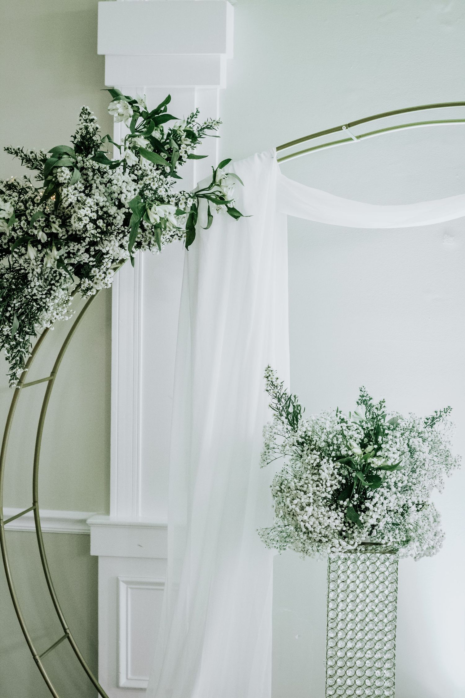 Elegant white floral arrangement with baby's breath in a crystal vase against a white column and sheer curtain backdrop.