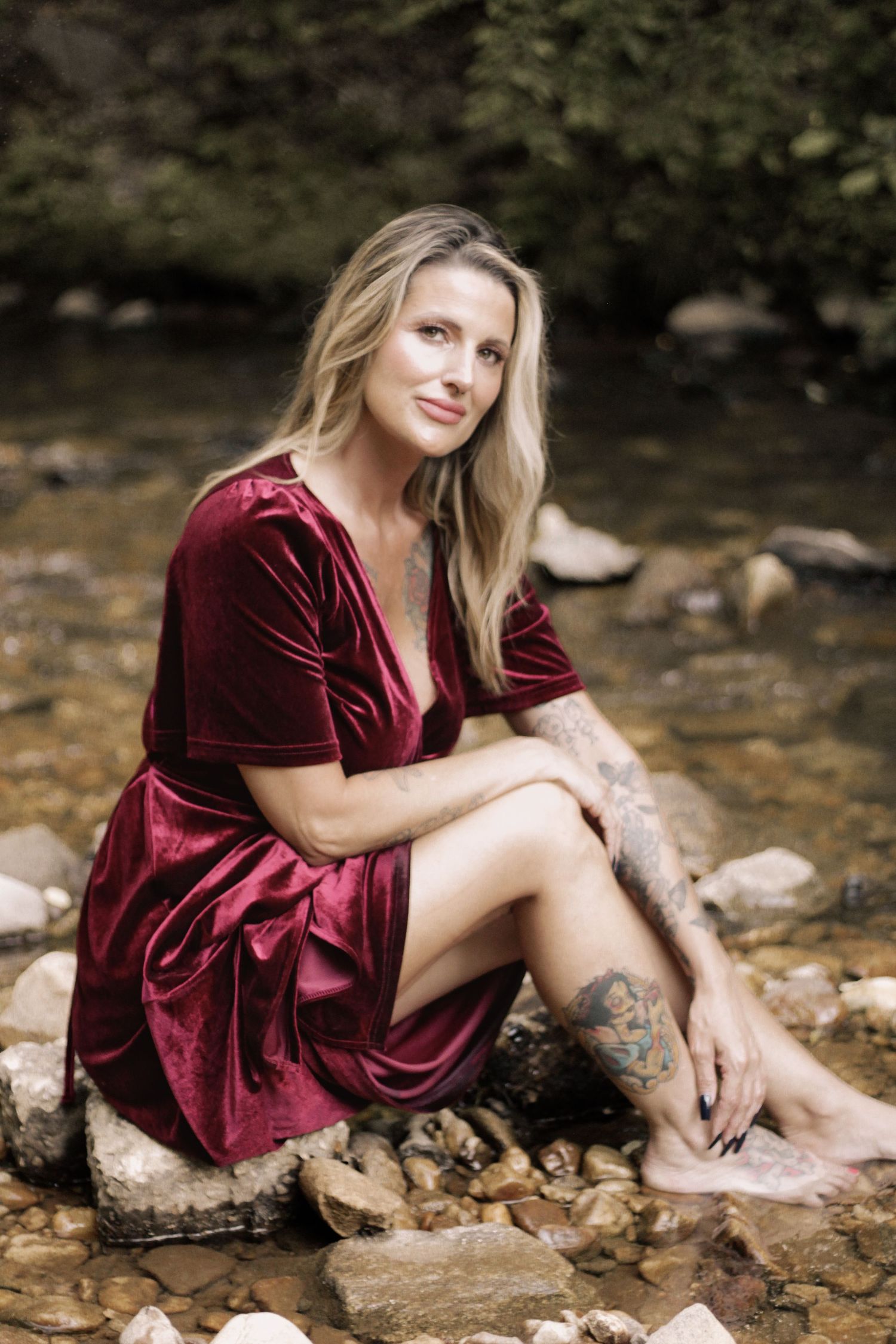 Woman in burgundy velvet dress poses gracefully by a stream with rocks in a natural outdoor setting.