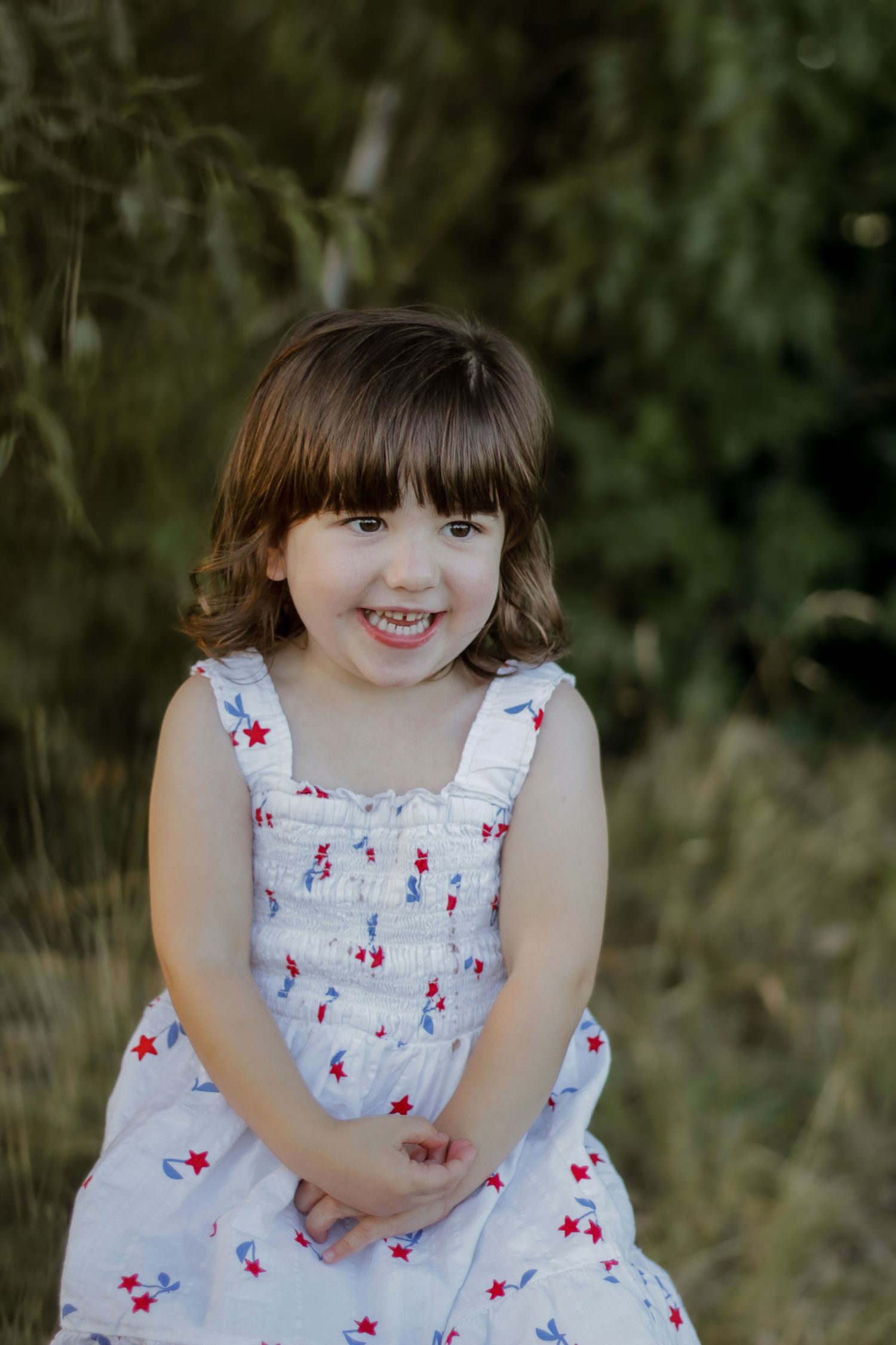 Small child wearing a white patterned dress smiles while sitting outdoors.