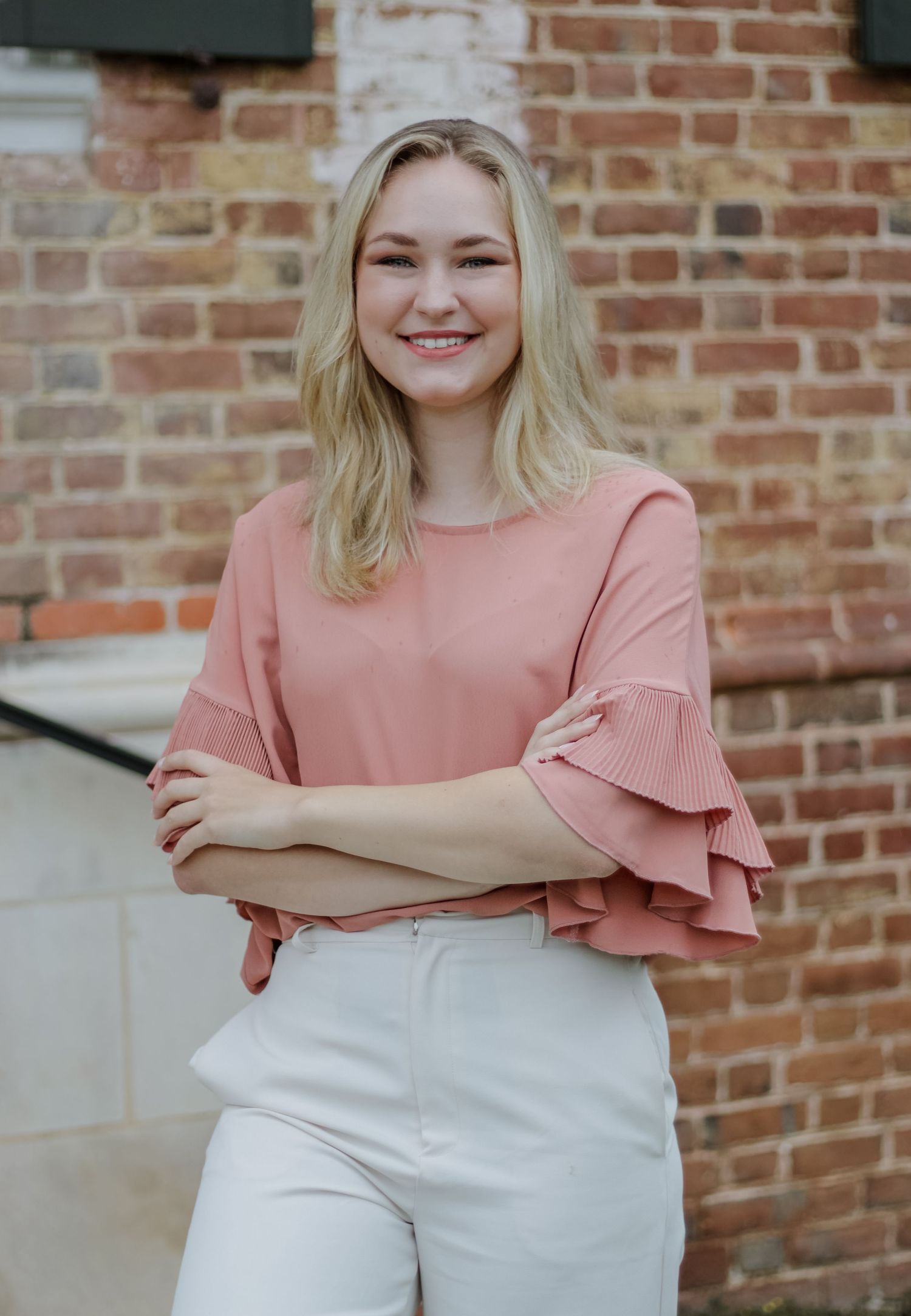 Professional portraits against brick wall featuring pink ruffled sleeve top and white pants.