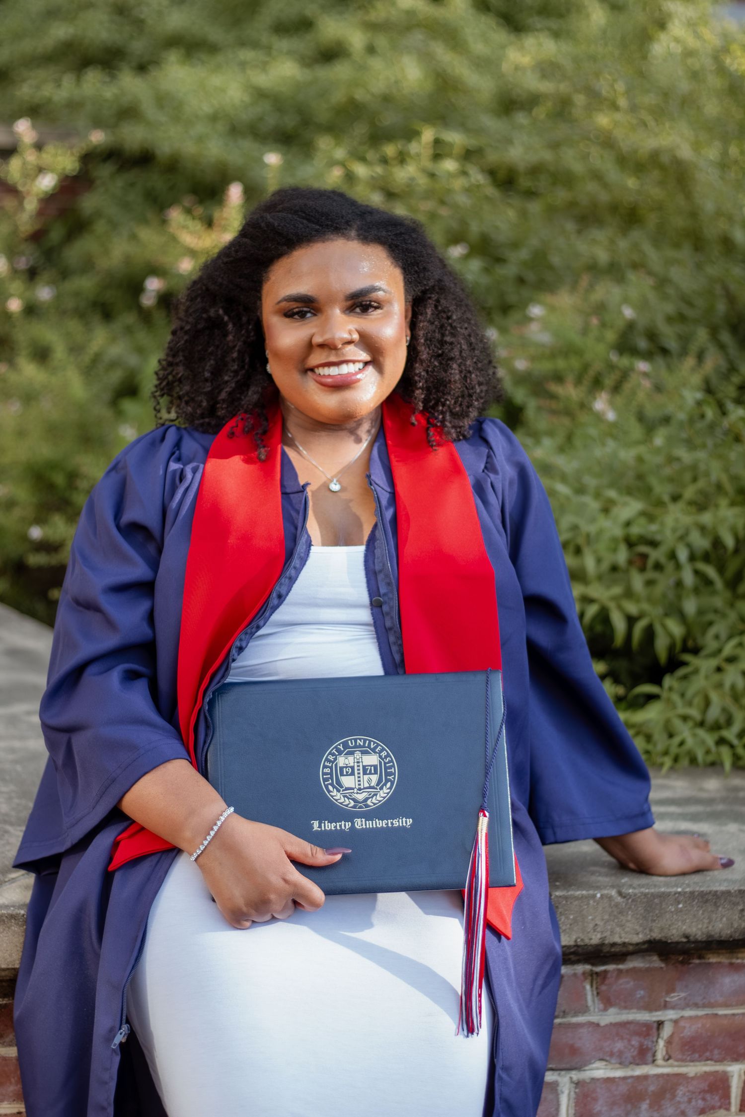 Graduate in navy blue cap and gown holding diploma with red stole.