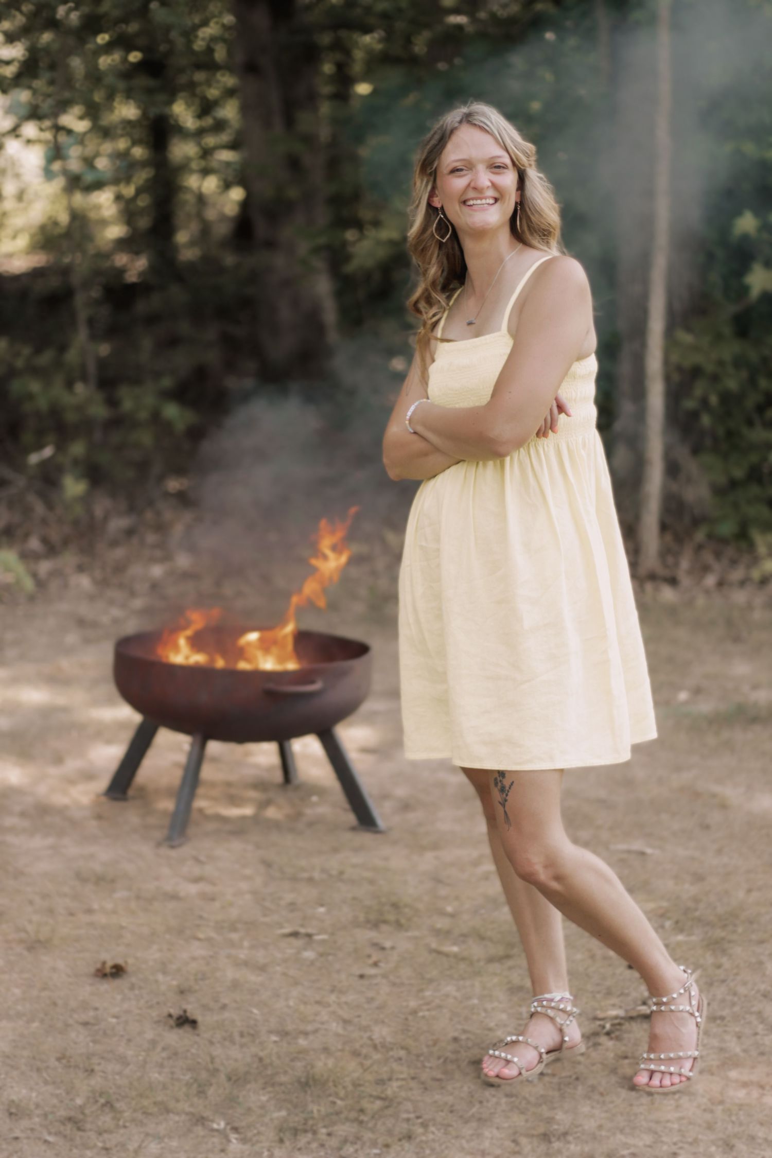 A person in a light sundress stands near a burning fire pit outdoors.
