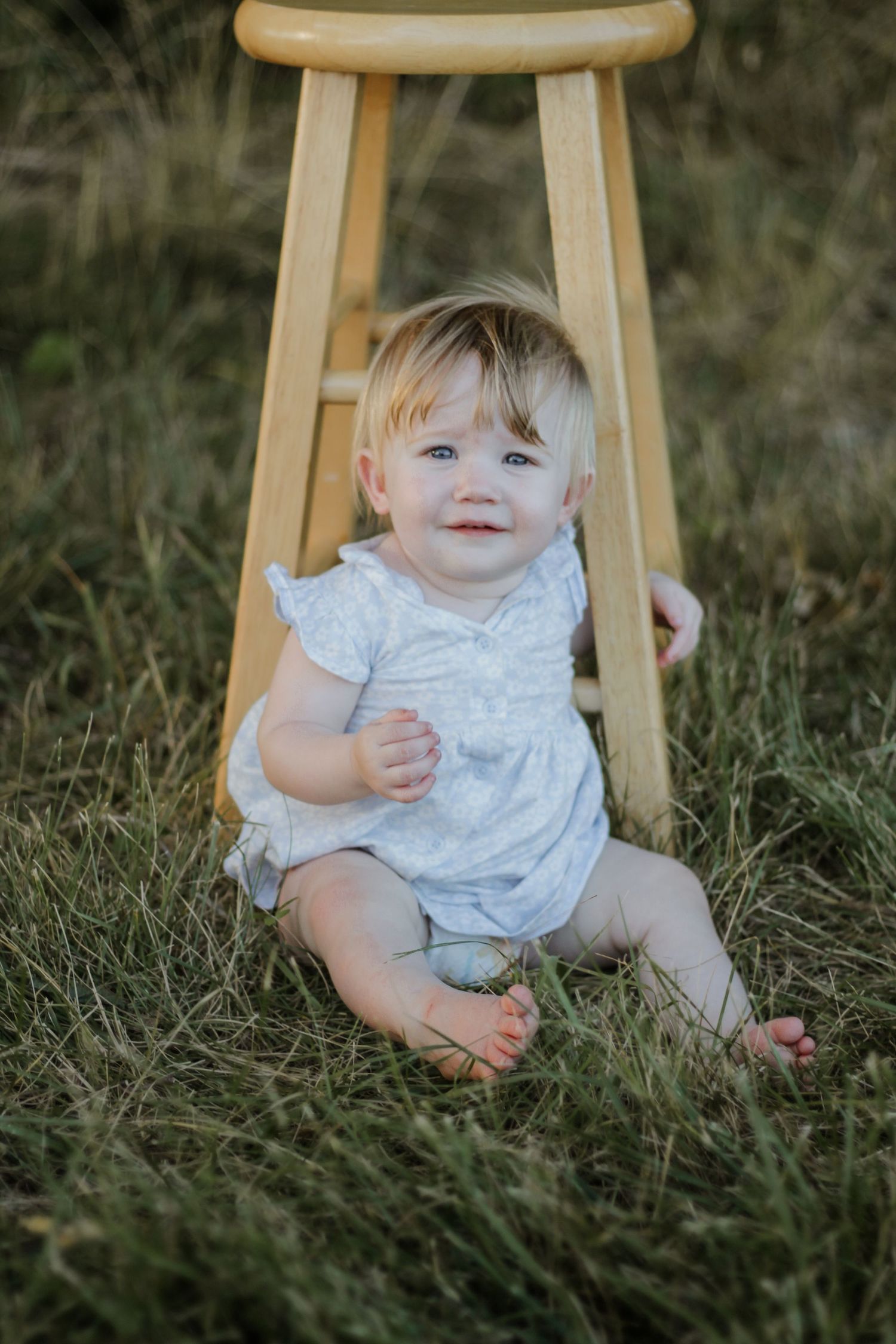 Baby in white dress sitting on wooden stool outdoors in grass.