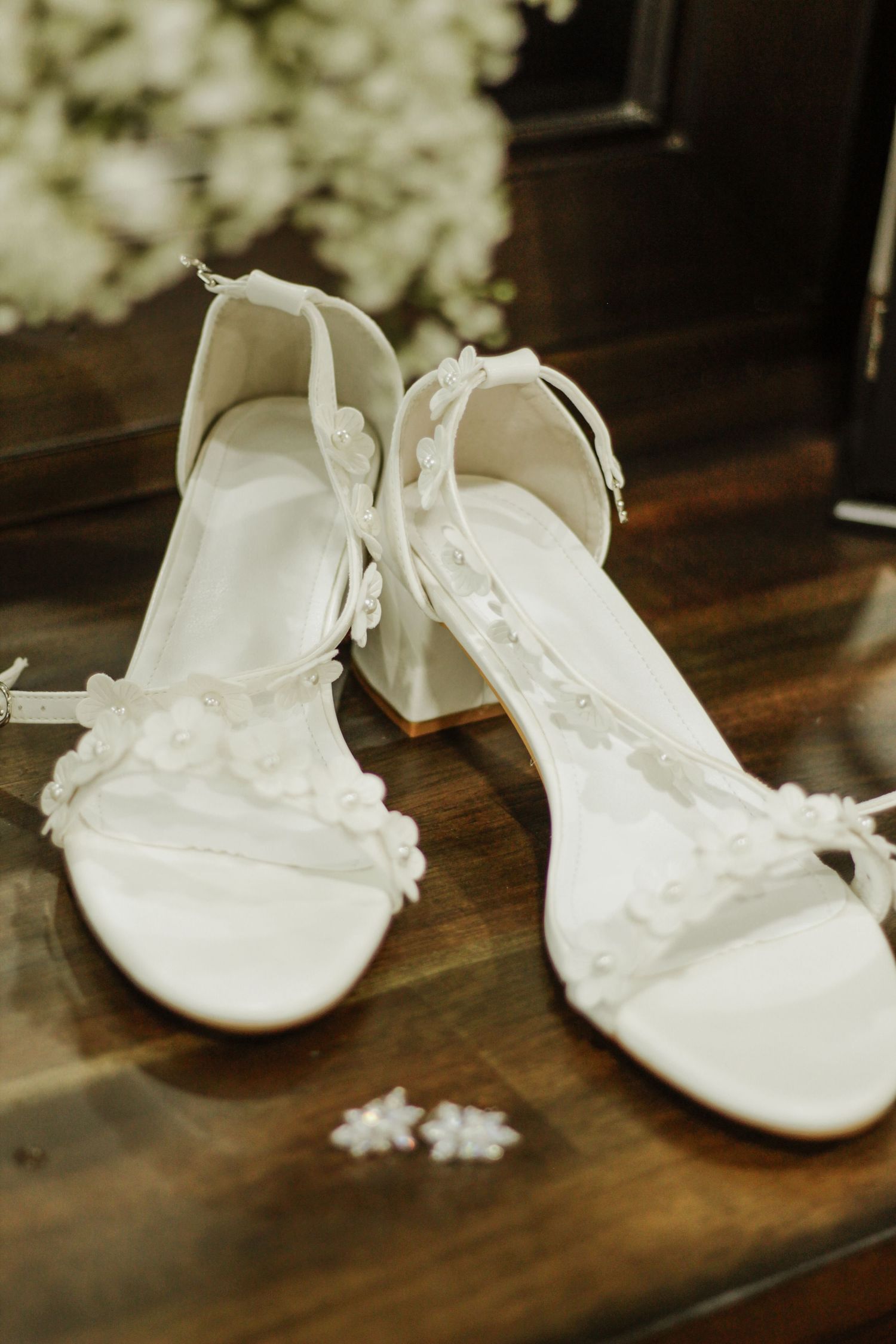 White wedding shoes on a dark wooden surface display delicate lace and floral details.