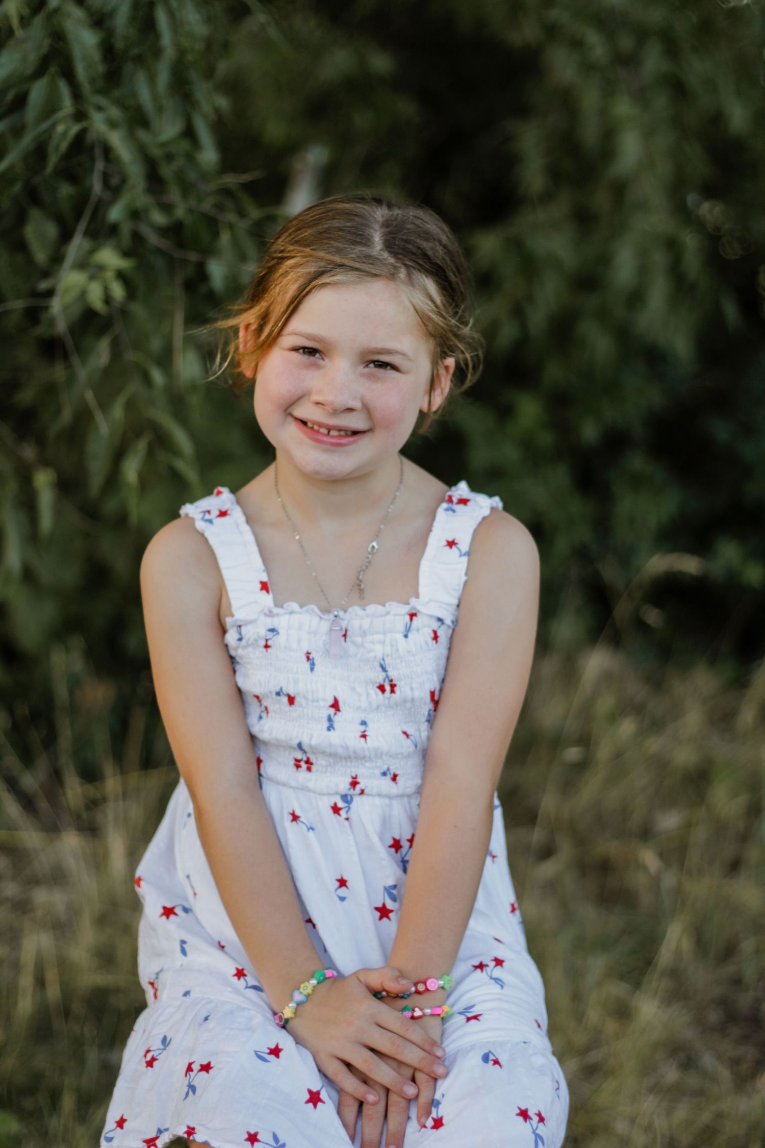 Young person in a white floral sundress poses outdoors against a green natural background.