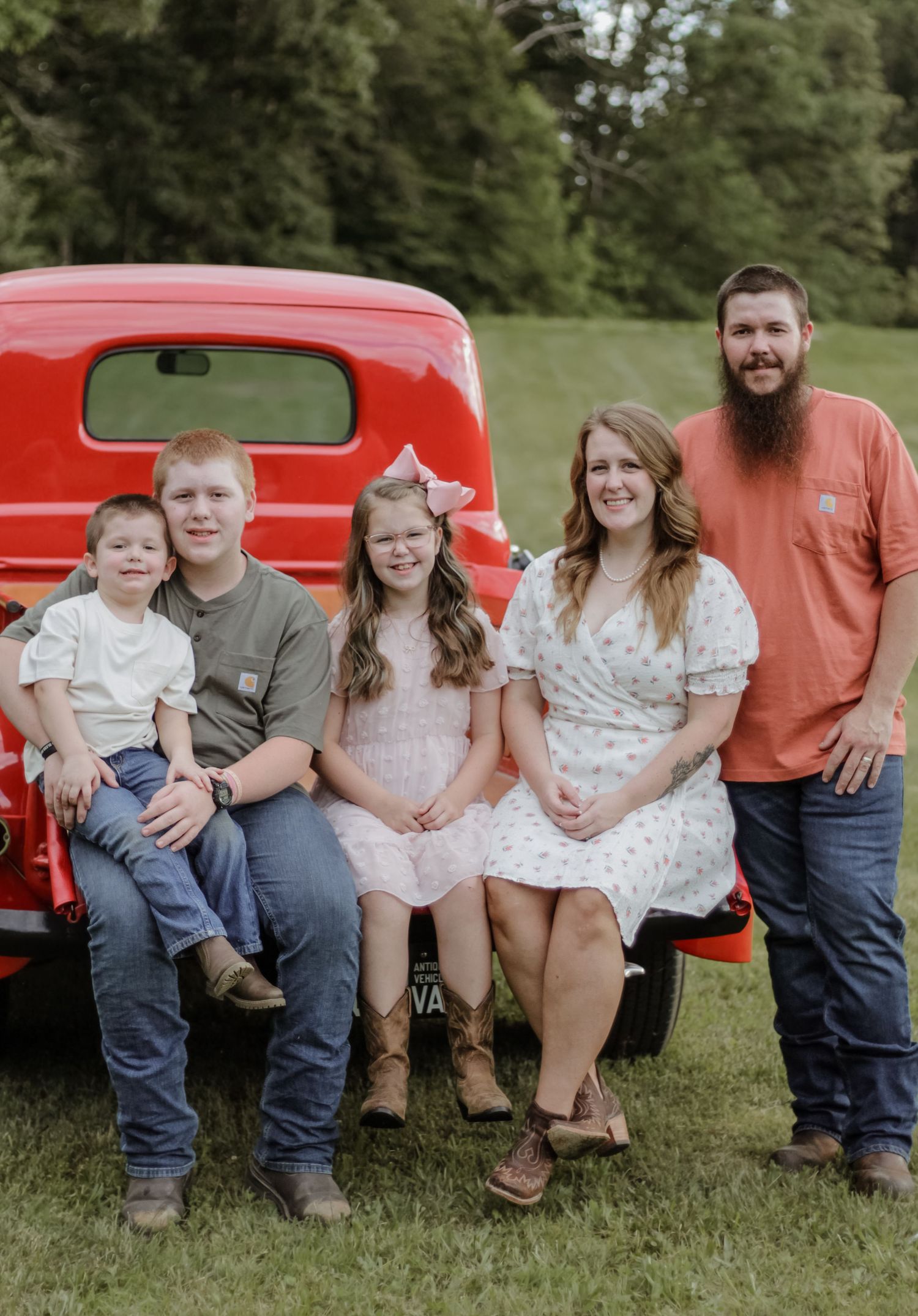 A casual family photo session sitting on a vintage red truck in a grassy outdoor setting.