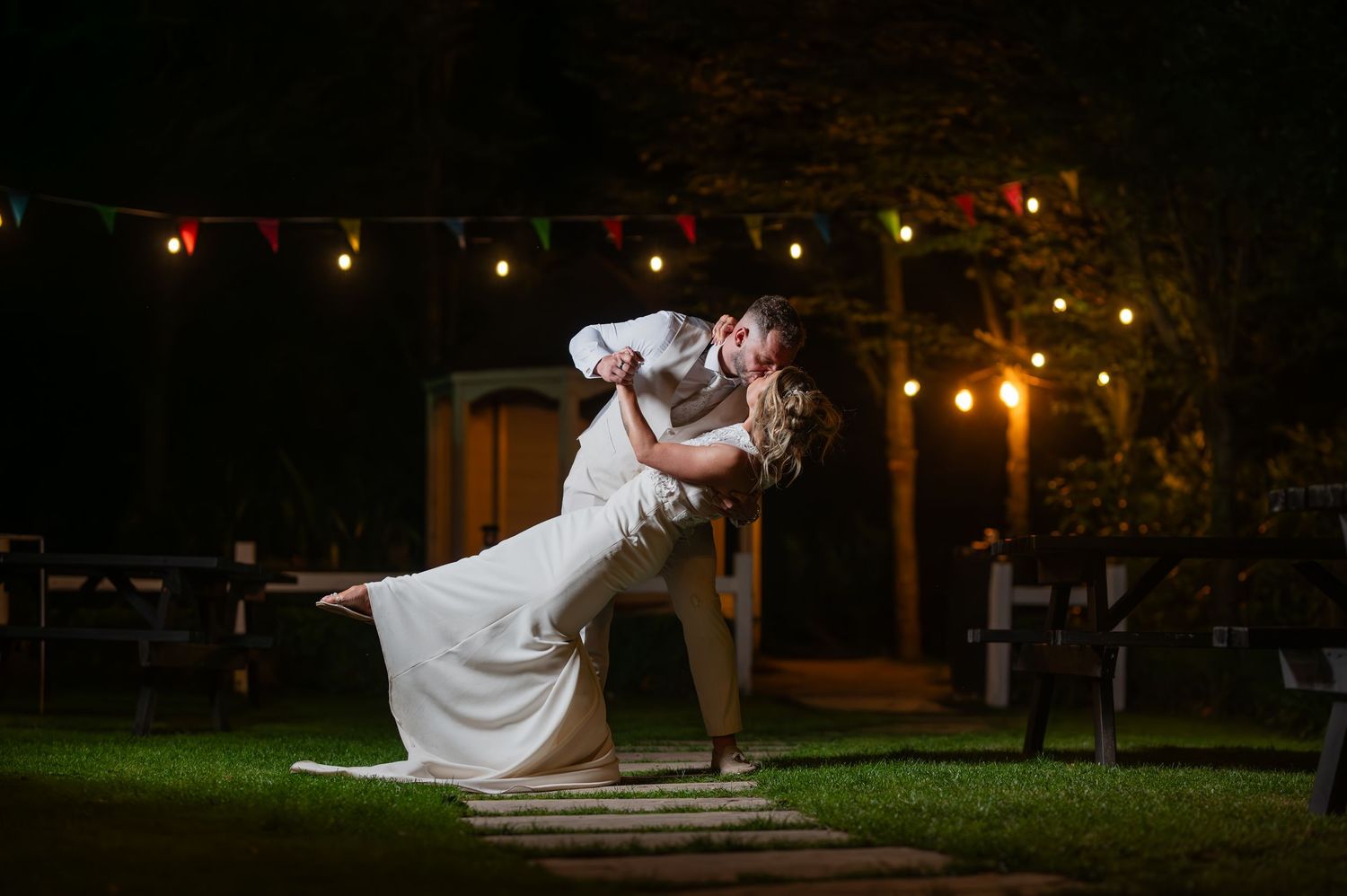 Romantic wedding dip kiss under string lights at night in outdoor garden reception venue.