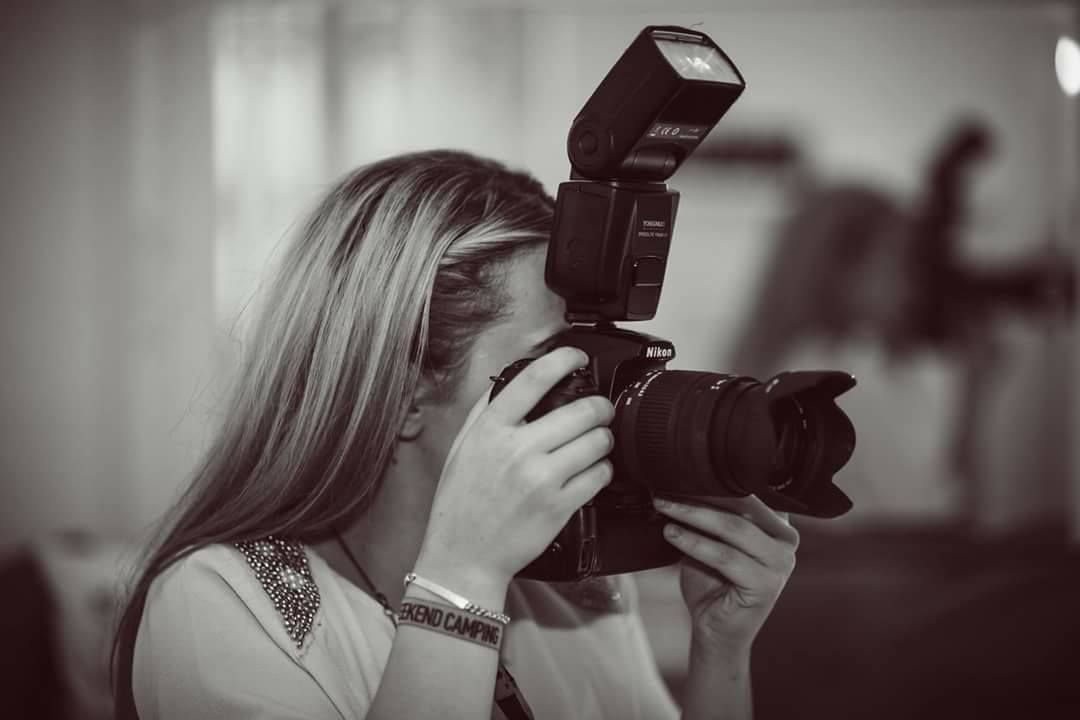 Black and white portrait of a female photographer holding a DSLR camera with flash attachment.