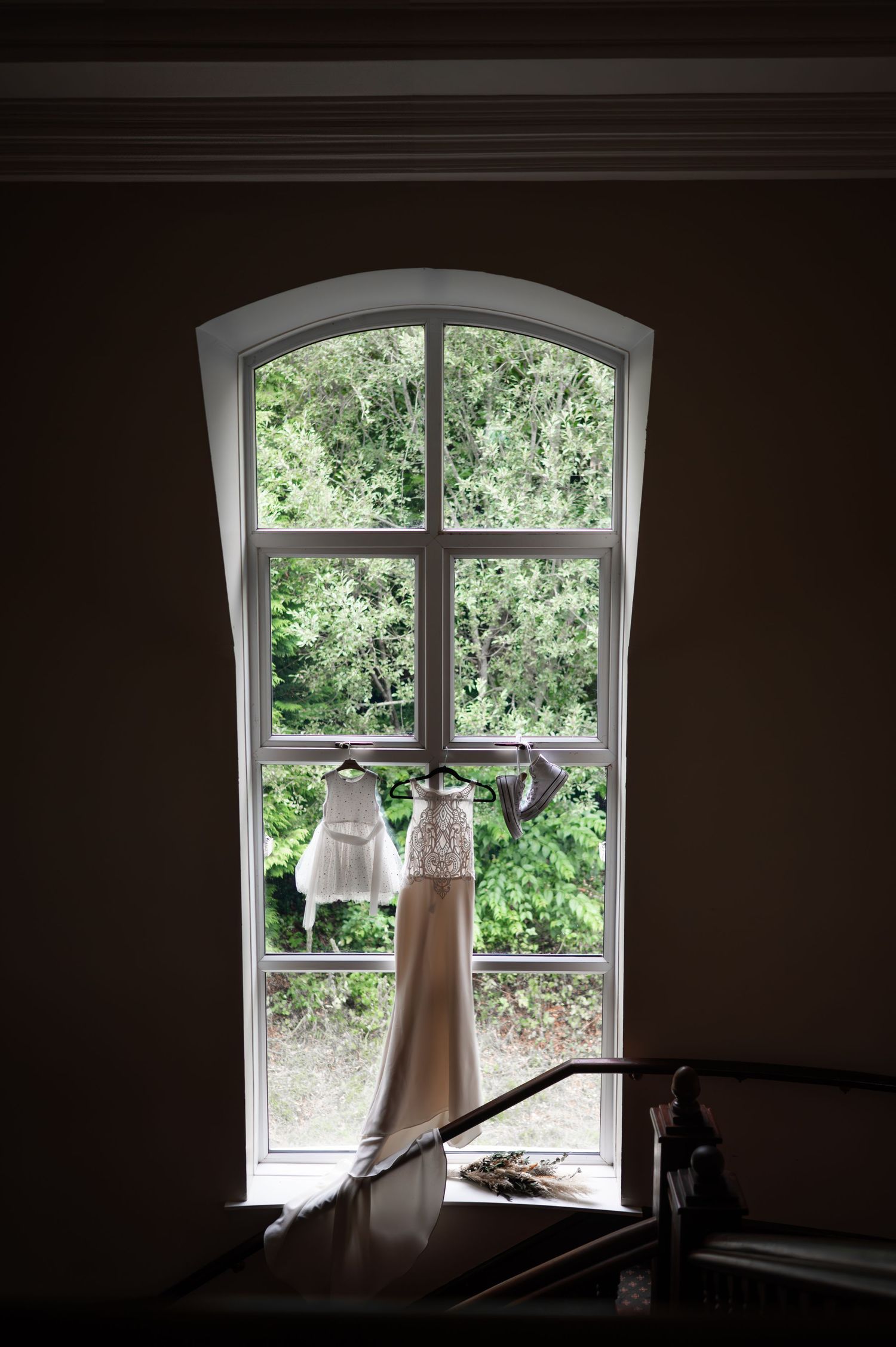 Wedding dress hanging in an arched window with lush green trees visible outside.