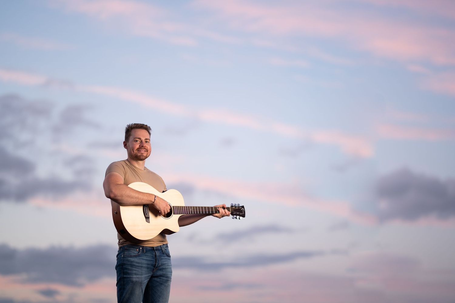 Man playing acoustic guitar outdoors against a pastel sunset sky with pink and blue clouds.