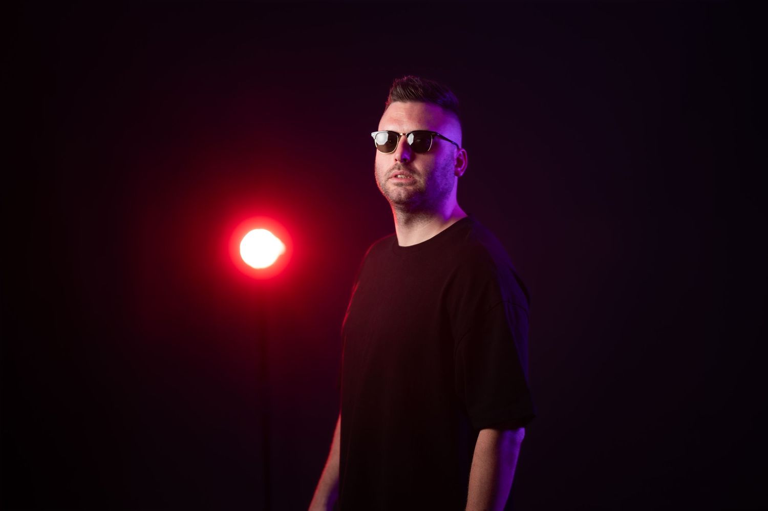 Man in black oversized t-shirt and sunglasses posing under dramatic red and purple stage lighting.