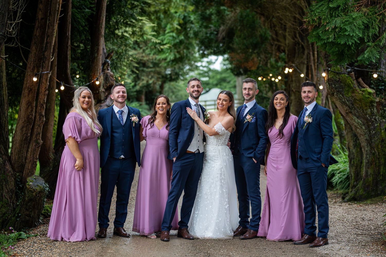 Wedding party posing outdoors among tall trees with string lights, bridesmaids in mauve gowns and groomsmen in navy suits.