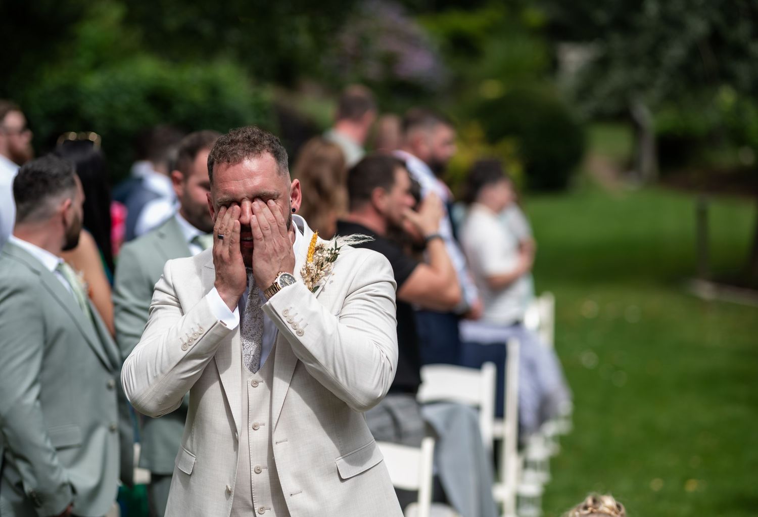 Emotional groom covers his face with hands during outdoor wedding ceremony while guests watch from white chairs.