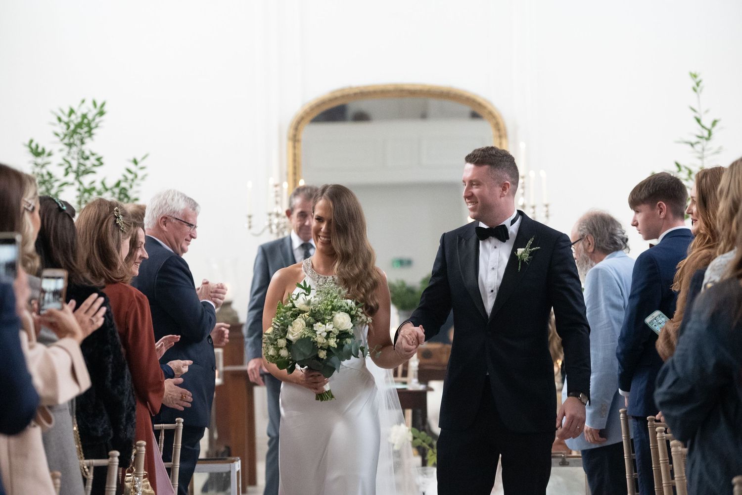 Bride and groom walk down the aisle smiling, bride holding white floral bouquet, guests applauding in elegant white venue.