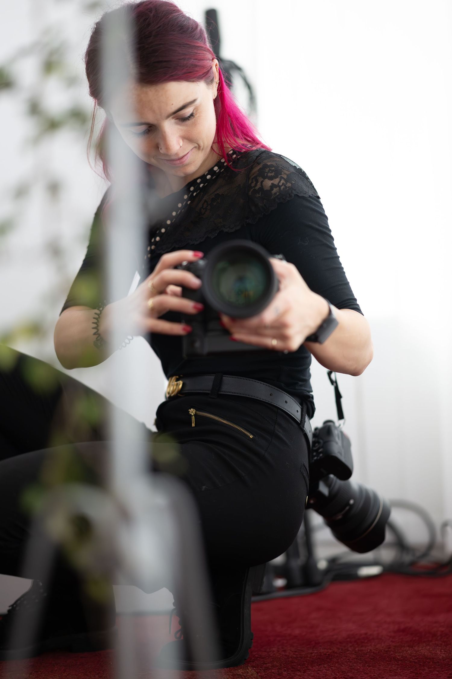 Pink-haired female photographer crouching while reviewing shots on professional DSLR camera indoors.