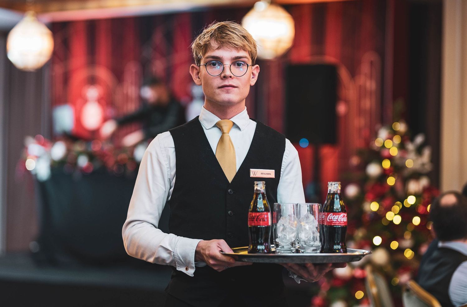 Young waiter in vest and tie carrying a tray with Coca-Cola bottles and glasses at an elegant event.