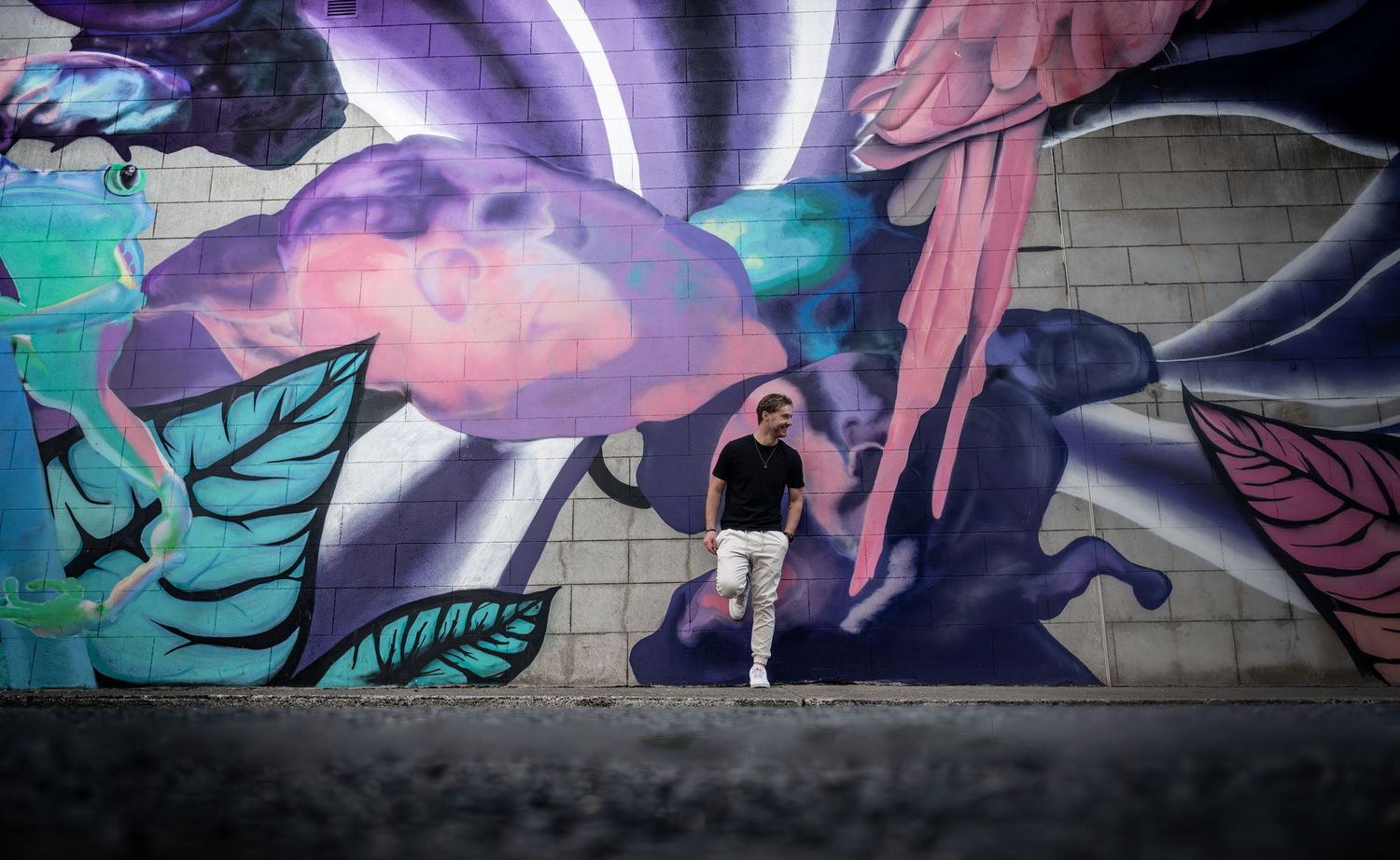 Young man in black outfit walking along a bold colorful street art mural in Dublin by a brick wall.