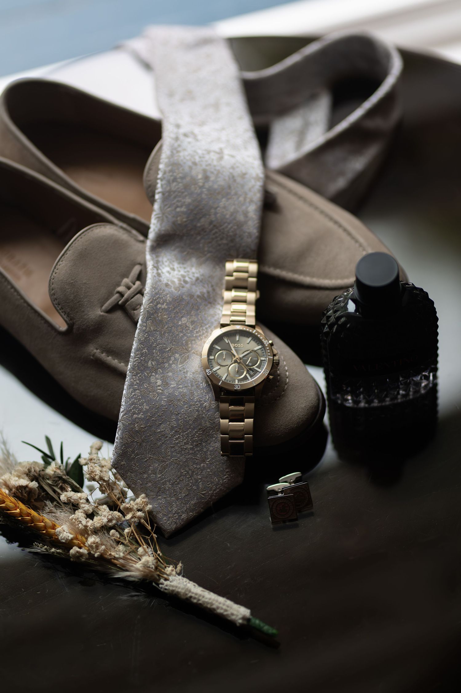 Groom wedding accessories including gold watch, grey tie, loafers, boutonniere, and cologne.
