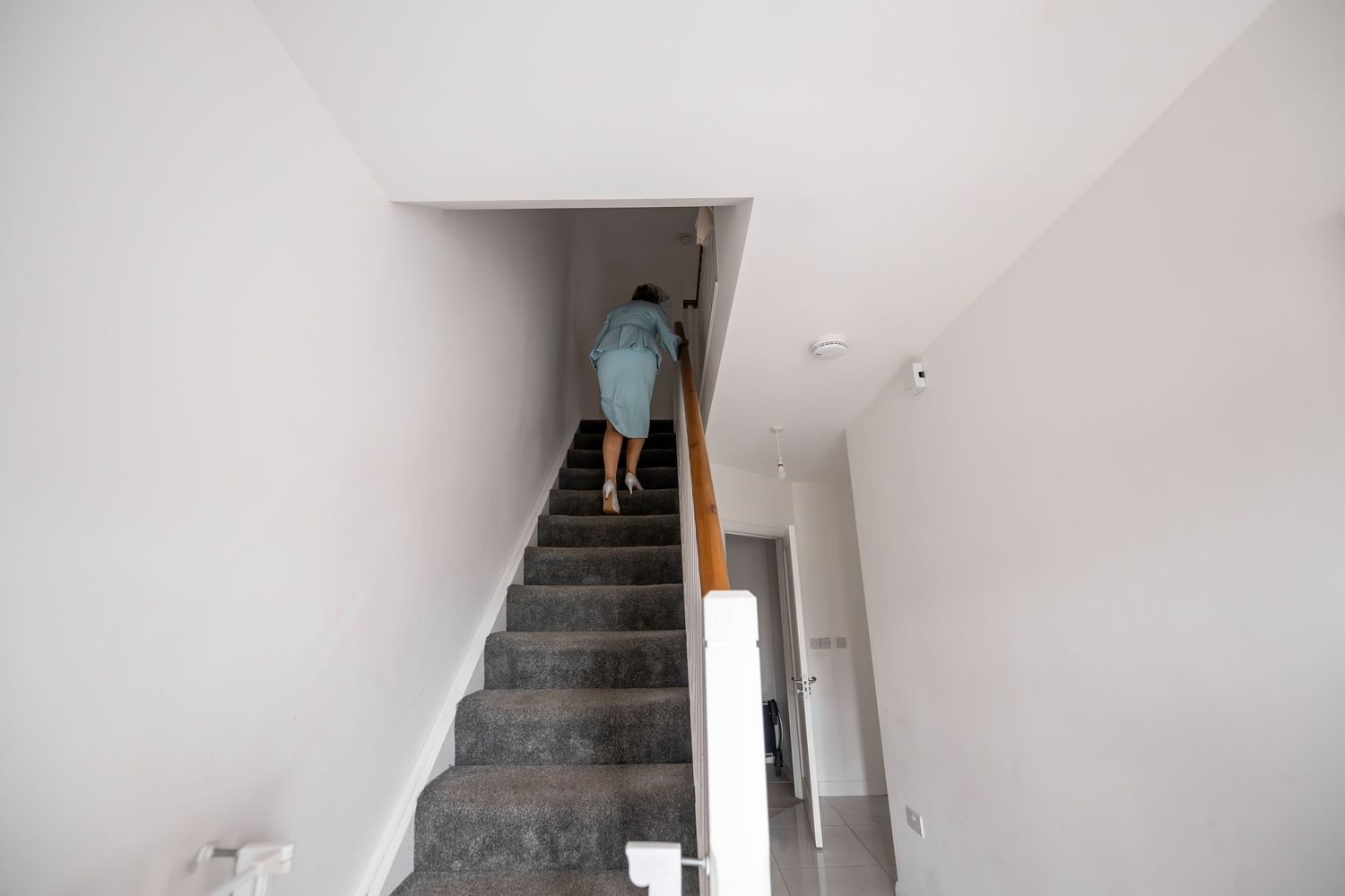Person walking up carpeted stairs with white walls and wooden handrail in a modern home interior.