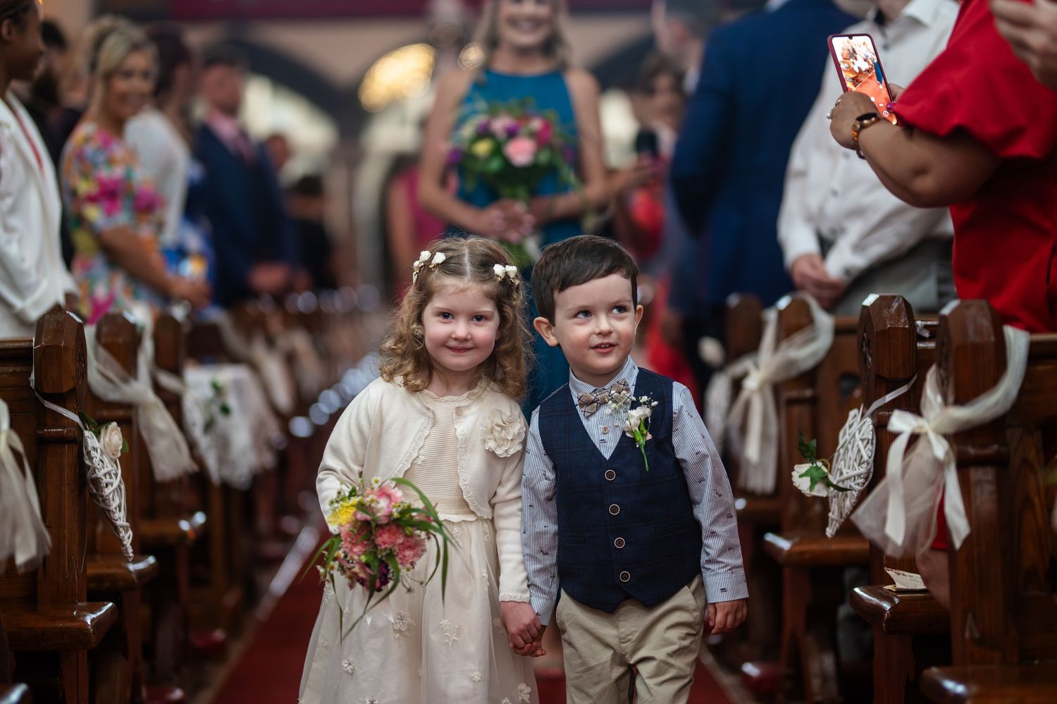 Flower girl and ring bearer walking down church aisle hand in hand at a wedding ceremony.
