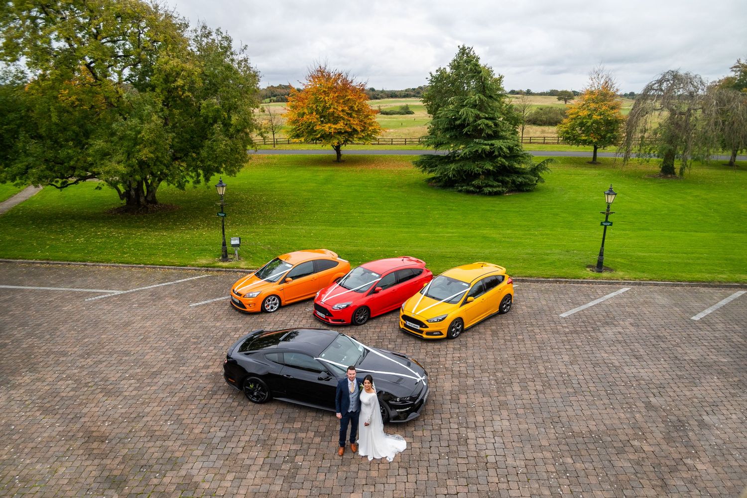Bride and groom pose beside four colorful Ford cars on a cobblestone driveway at a scenic countryside wedding venue.