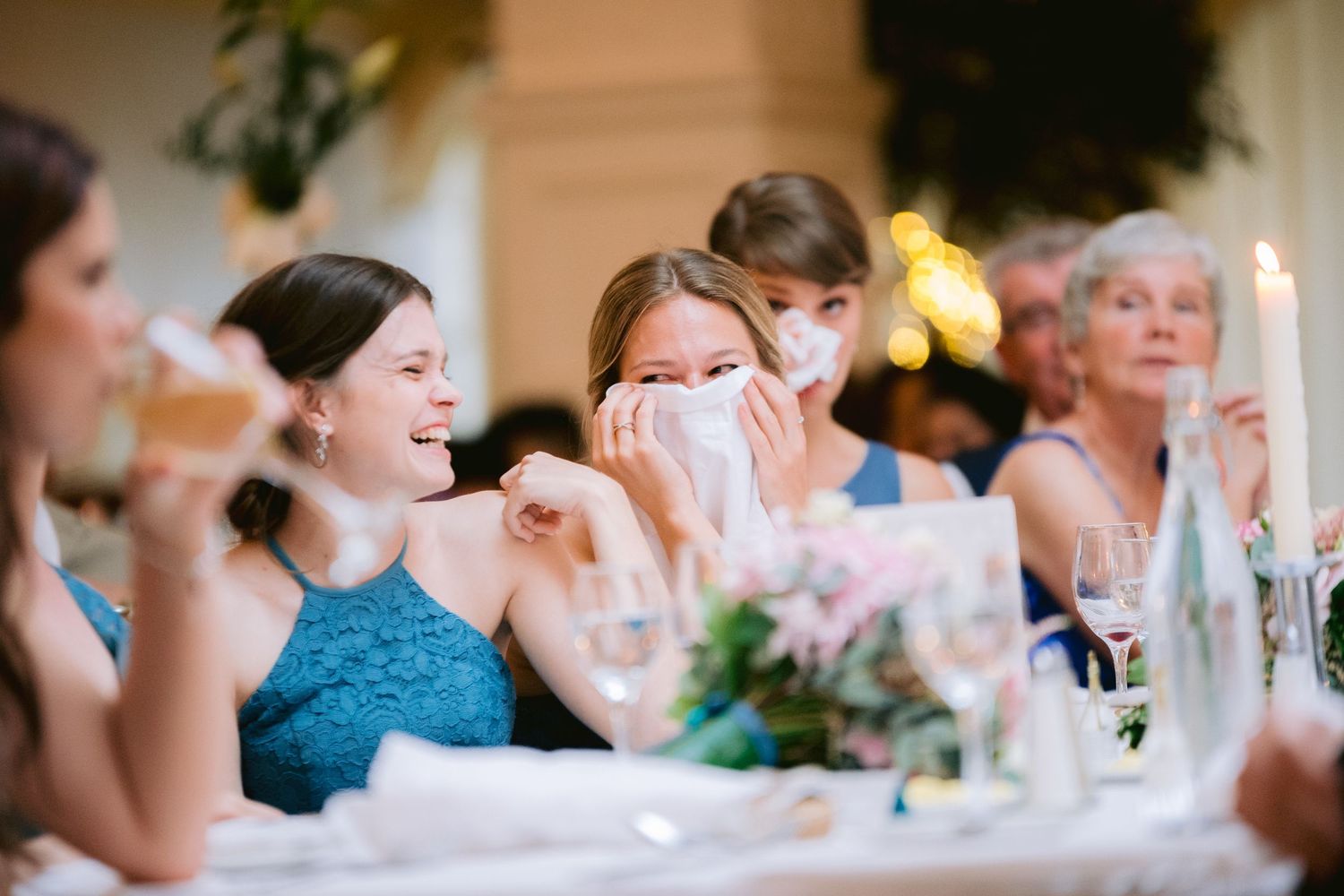 Bridesmaids in blue dresses laugh and cry with emotion during wedding reception speeches at elegant venue.