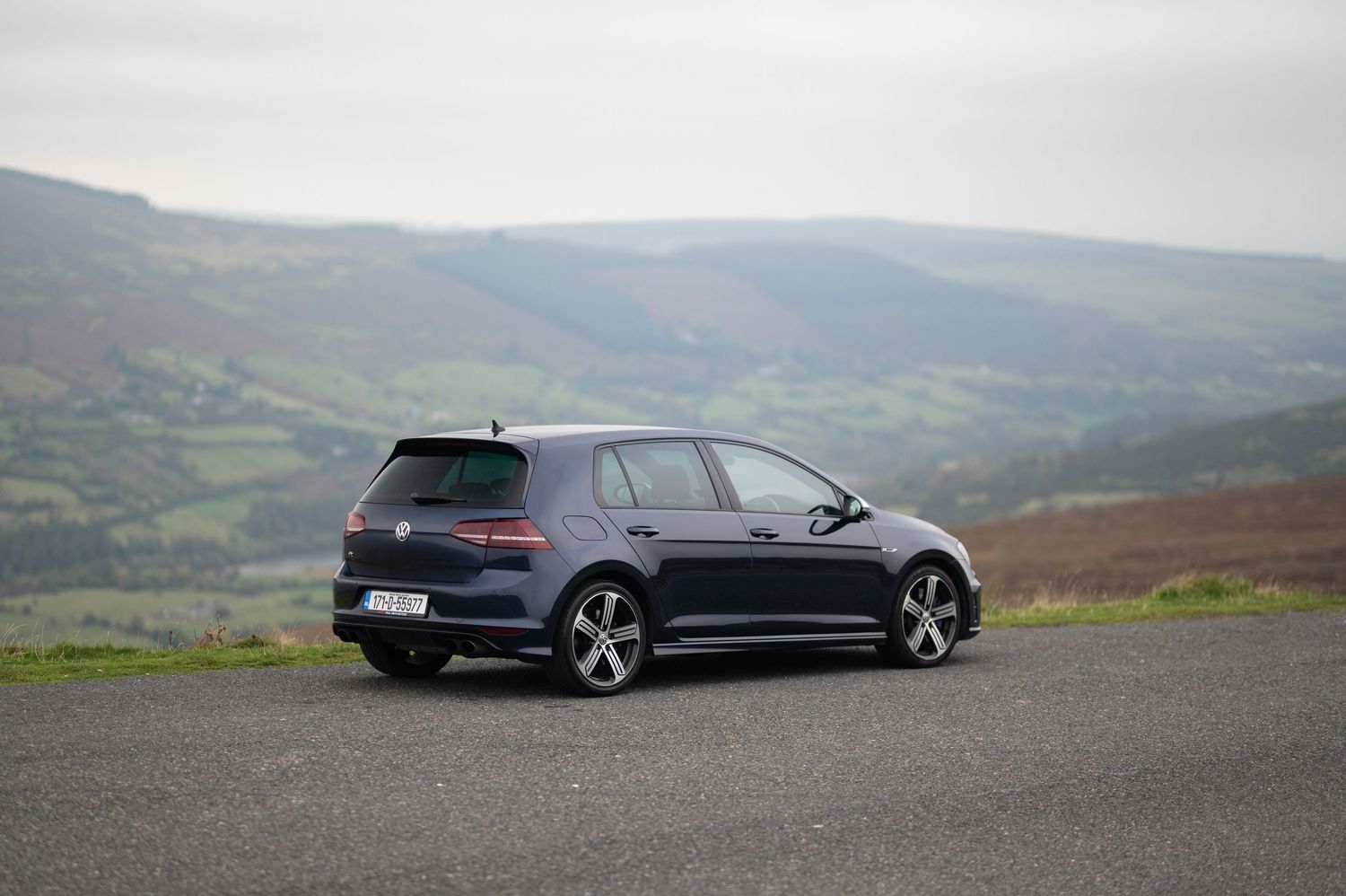Navy blue Volkswagen Golf R parked on a hilltop road overlooking a lush green valley.