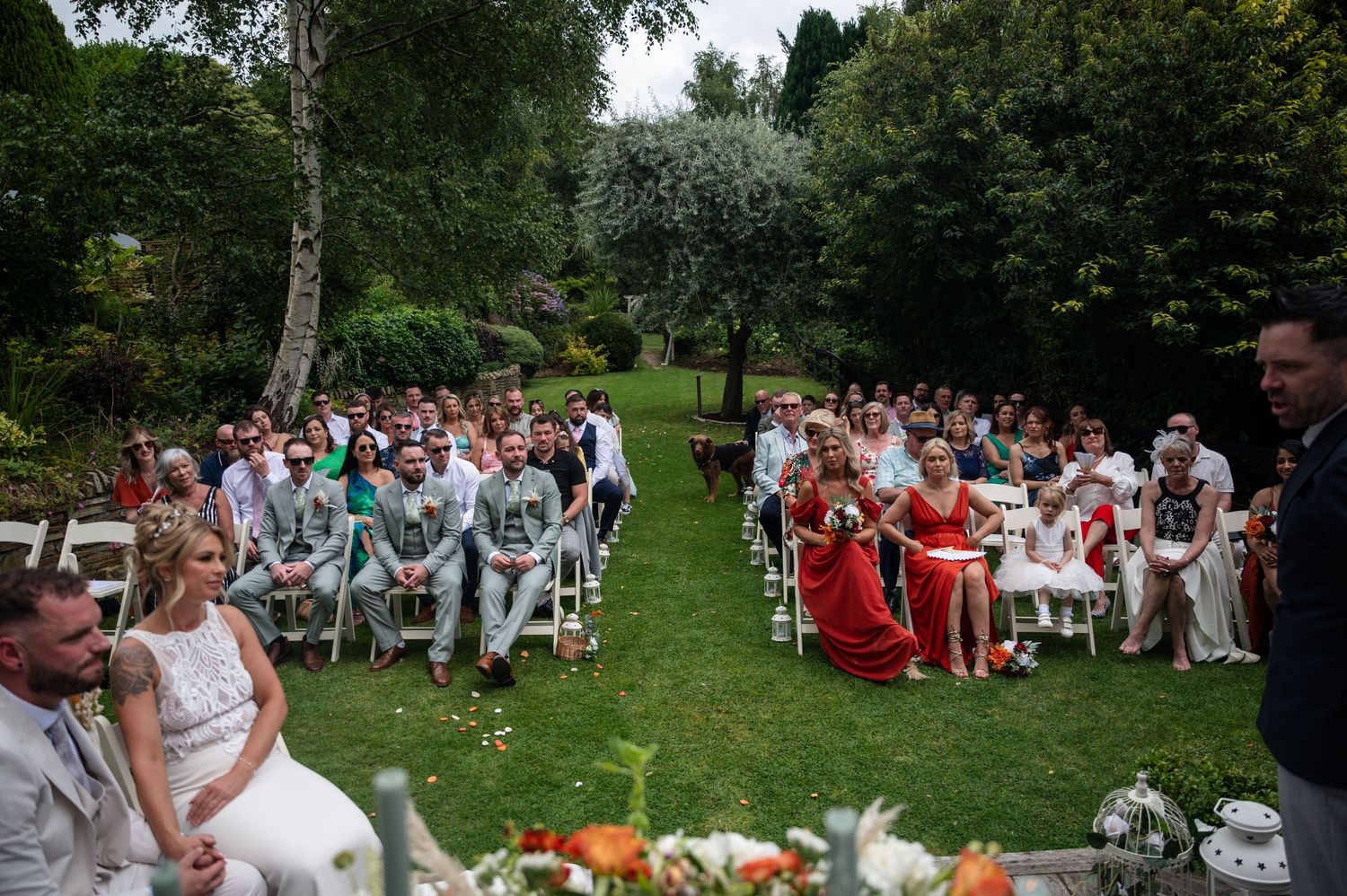 Wide angle view of outdoor garden wedding ceremony with guests seated on white chairs in lush greenery.
