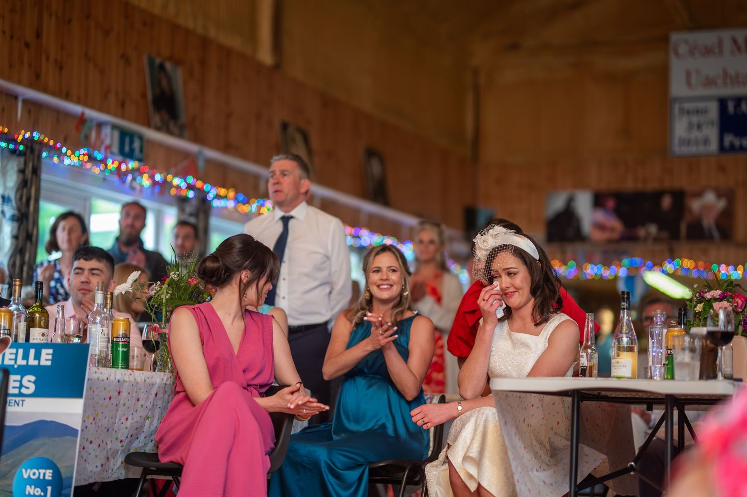 Women in formal attire laugh and clap at an indoor event, seated at a table with drinks in a decorated hall.
