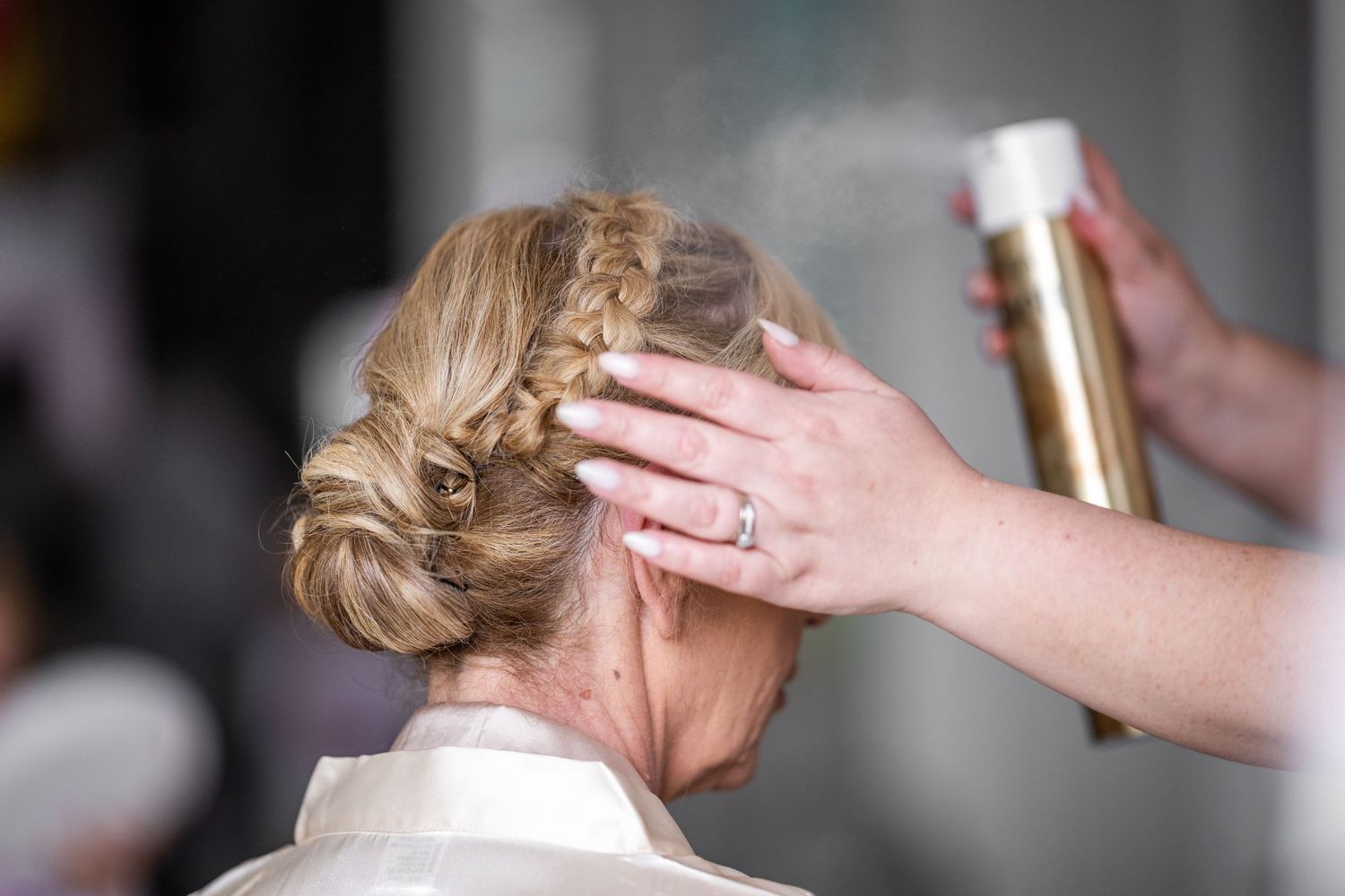 Hairstylist applying hairspray to an elegant braided updo on a blonde woman at a salon.
